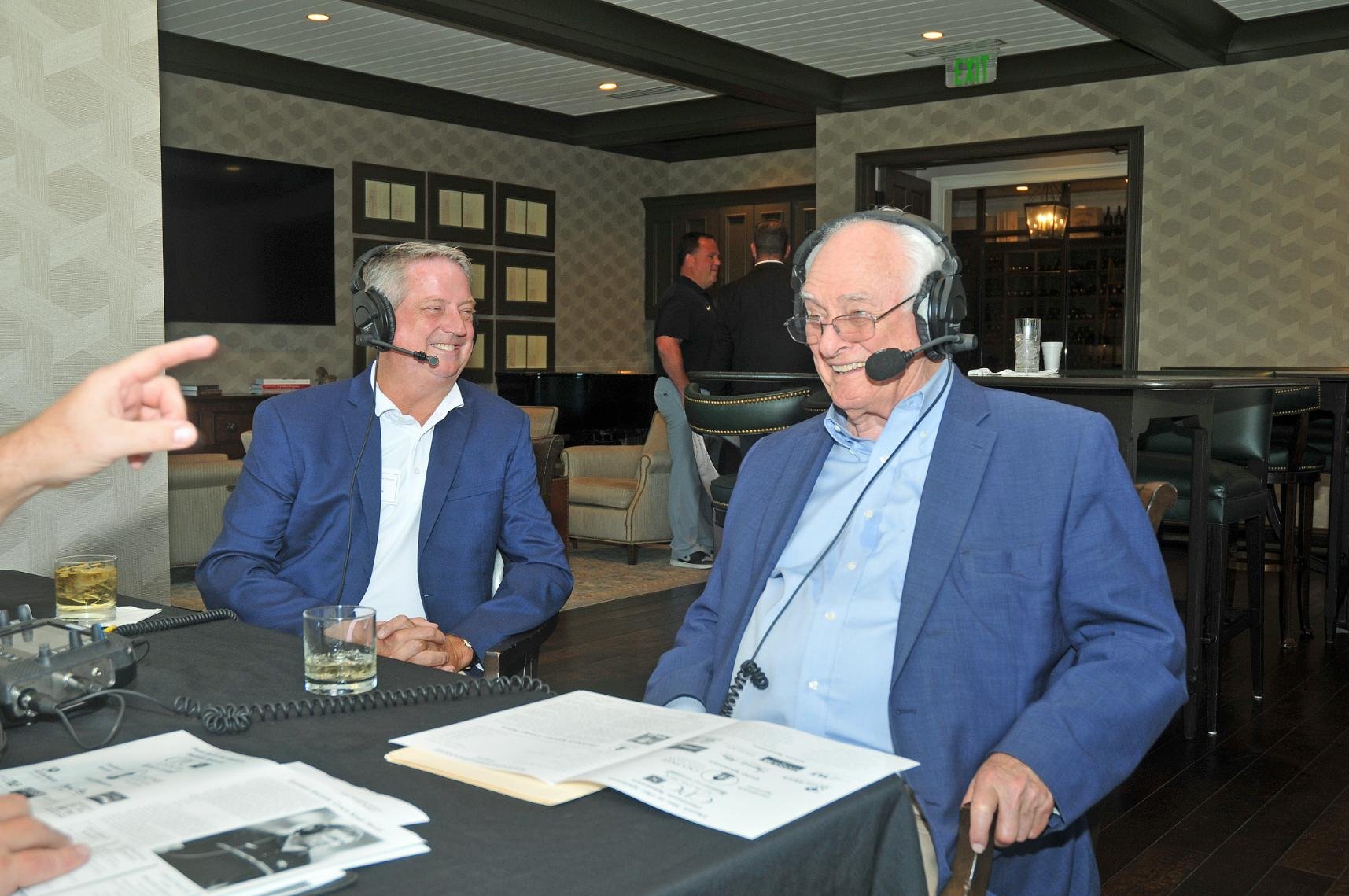 Two men wearing headsets and suits sitting at a table with papers and a recording device, engaged in a conversation in a room with people in the background.