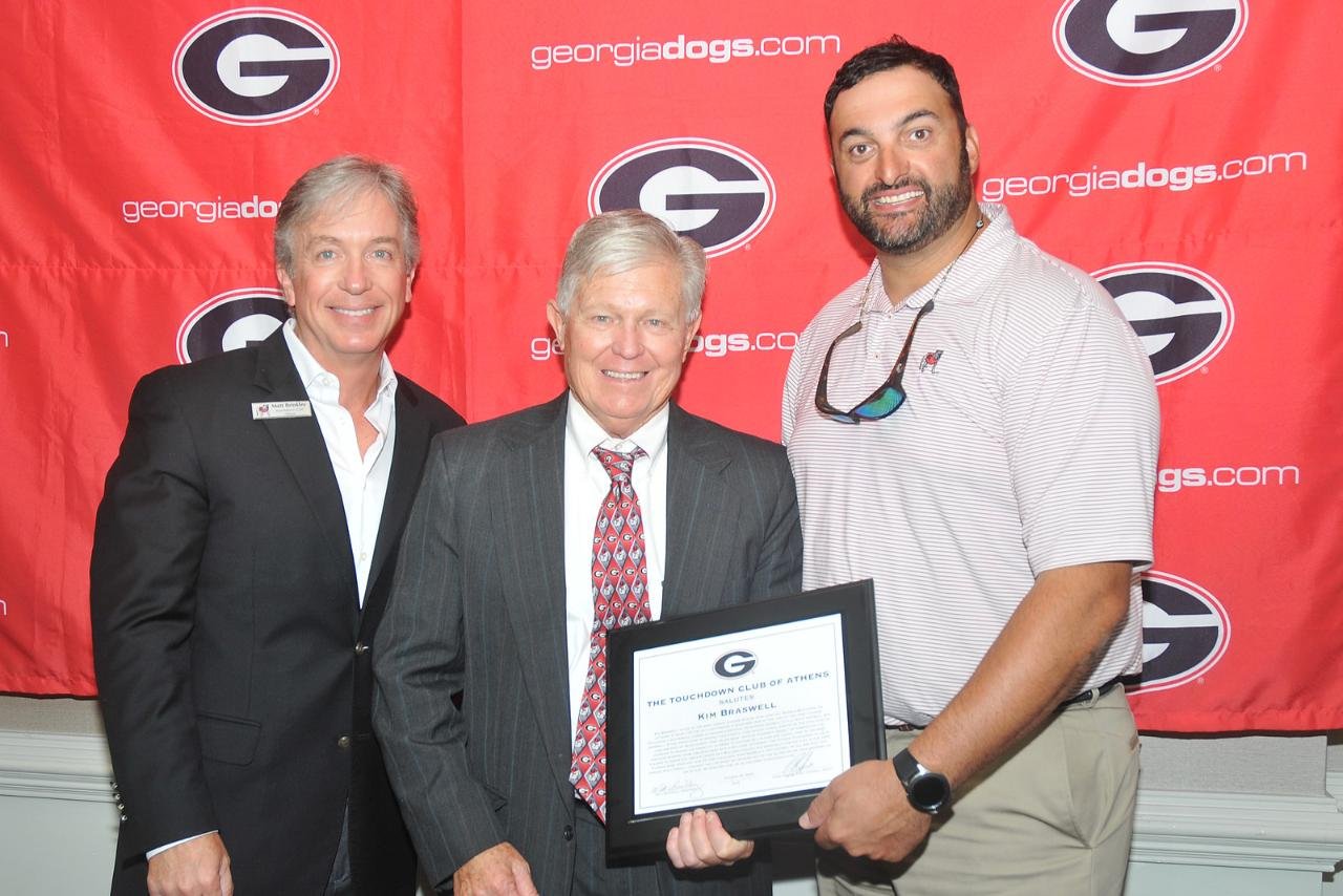 Three men posing in front of a Georgia Bulldogs backdrop, with one holding a framed certificate.