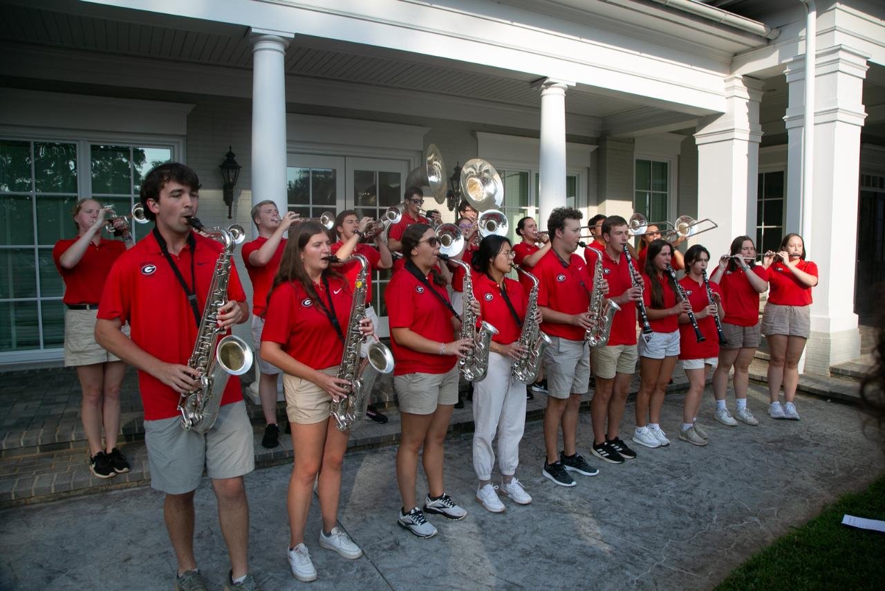 A marching band performing outdoors, wearing red shirts and khaki shorts. They play various instruments, including saxophones, trumpets, and sousaphones, standing in front of a building with white columns.