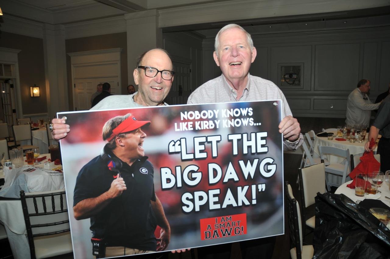 Two men holding a poster featuring a football coach with text "Nobody knows like Kirby knows... Let the Big Dawg speak!" at an indoor event.