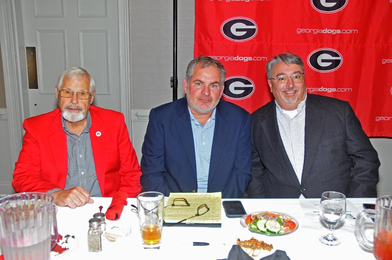 Three men sitting at a table with a red Georgia Bulldogs banner in the background, one wearing a red blazer, another in a blue blazer, and the third in a gray blazer, with food and drinks on the table.