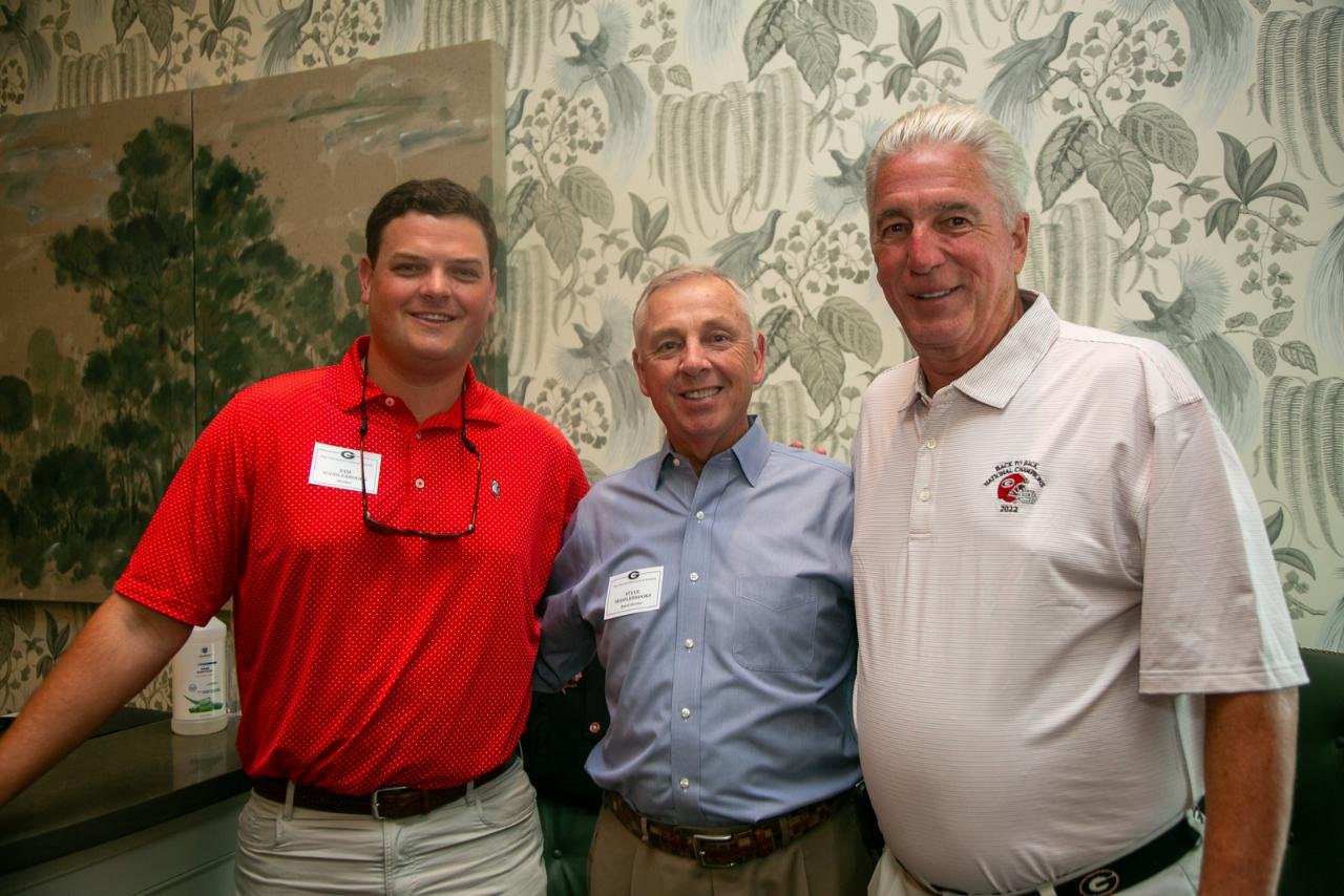 Three men posing together, one in a red polo shirt, another in a blue shirt, and the third in a white shirt. They are in front of a decorative wall with leafy patterns.