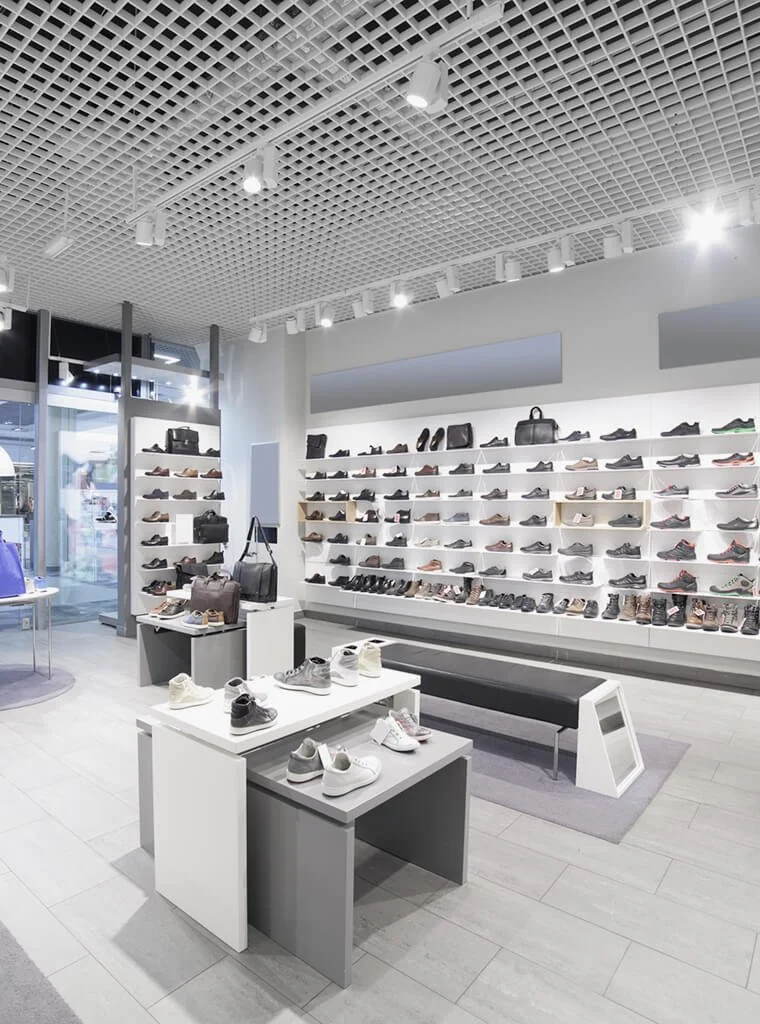 Interior of a shoe store with displays of various sneakers and dress shoes, handbags, and accessories on white shelves and tables, with modern lighting and a bench for customers to sit.