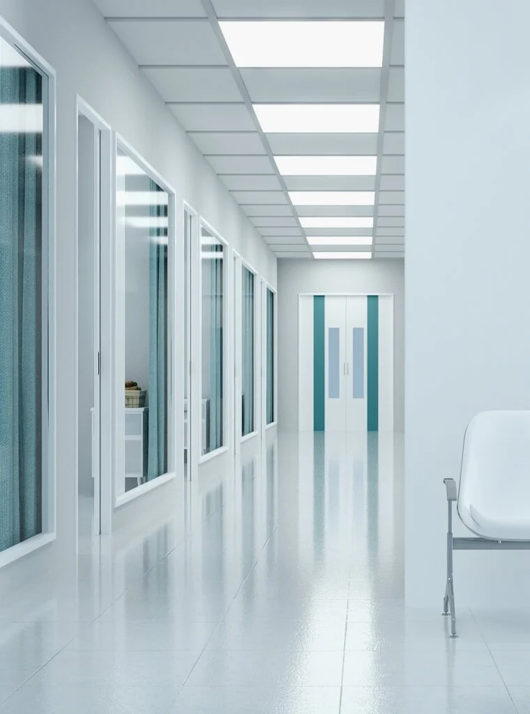 A clean, modern hospital corridor with glass doors on the left and a white chair on the right, leading to an elevator at the end.