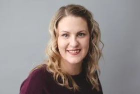 Portrait of a young woman with blonde curly hair, smiling, wearing a maroon top against a plain gray background.