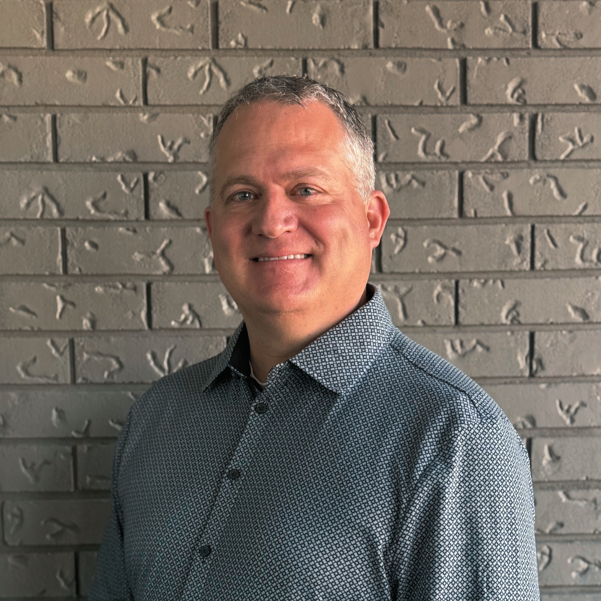 A smiling man wearing a patterned shirt standing in front of a gray brick wall.