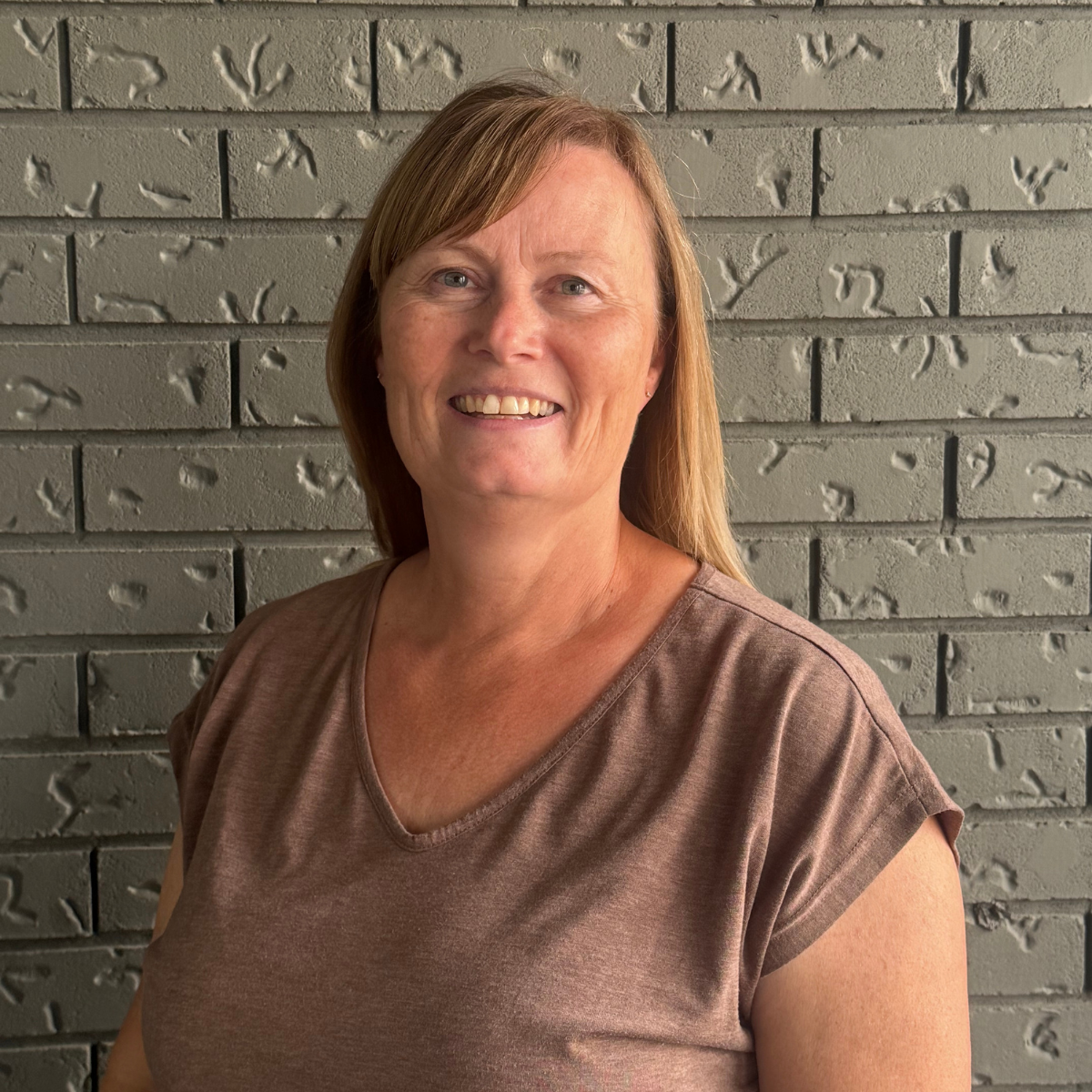 A smiling woman with long hair stands in front of a gray brick wall.