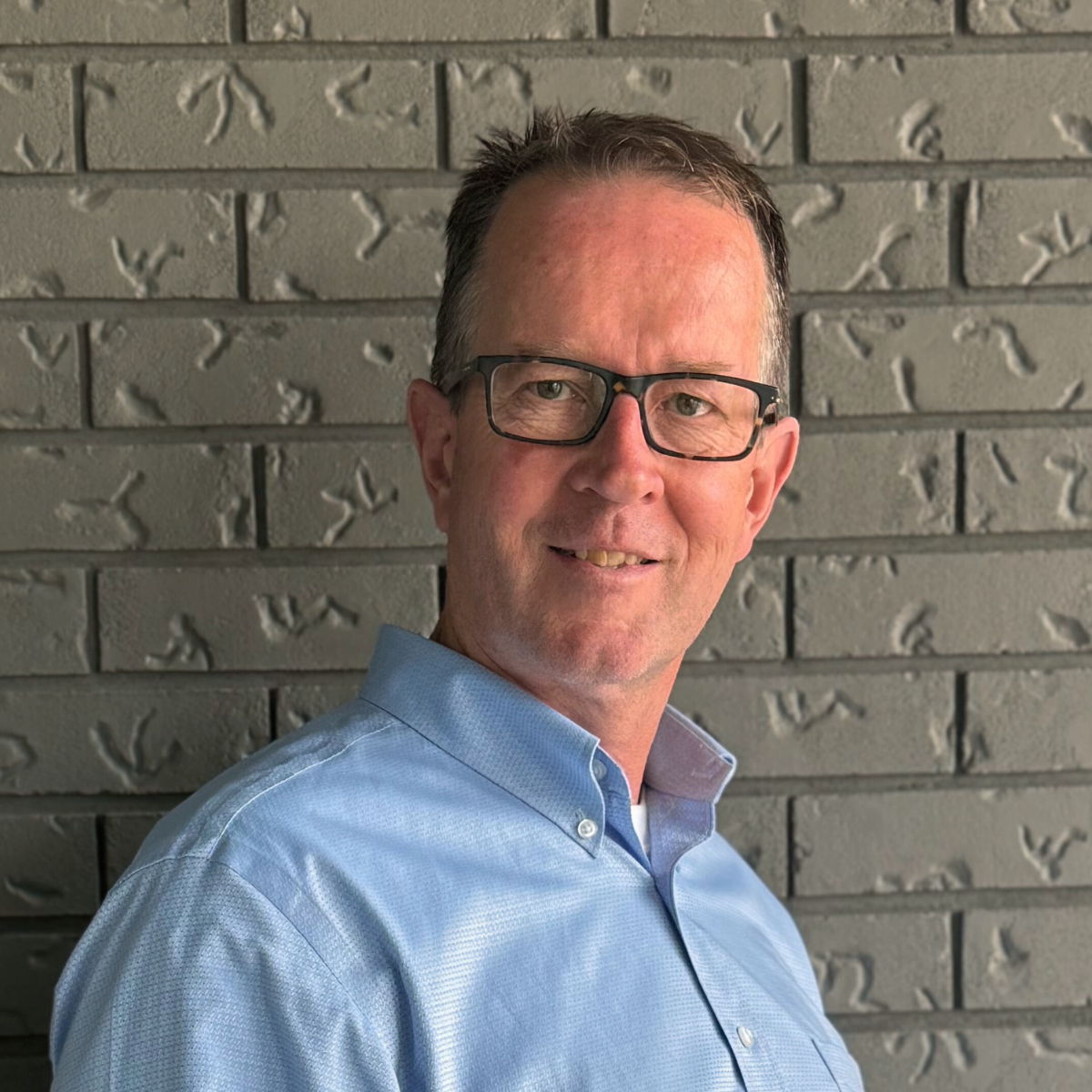 Headshot of a middle-aged man with glasses in a light blue button-up shirt, standing against a gray brick wall, smiling.