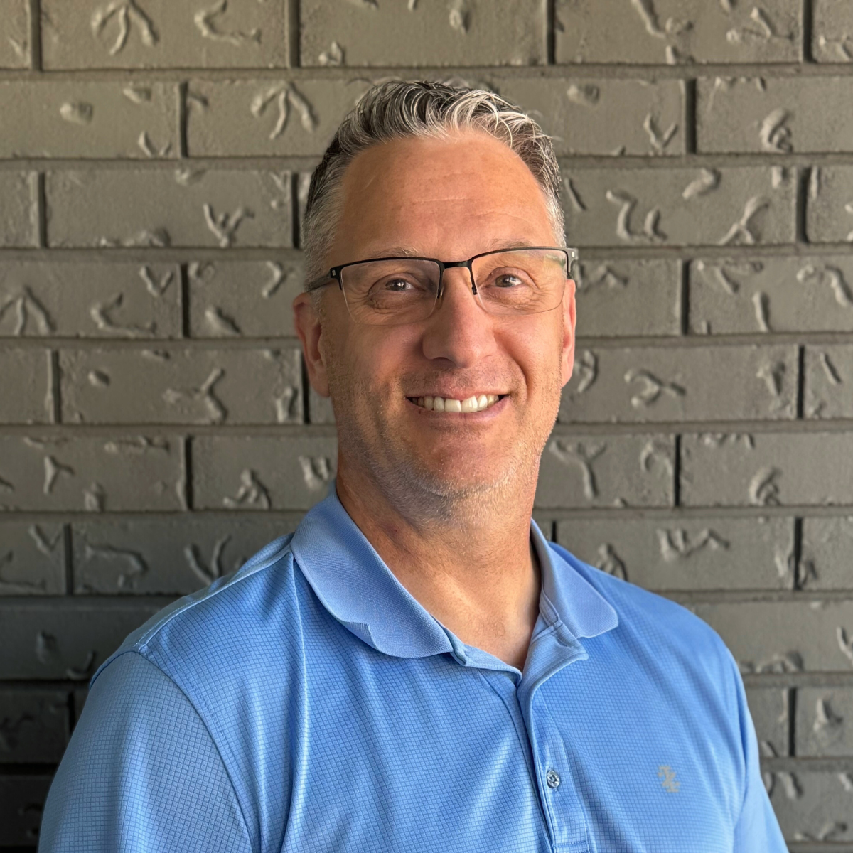 A man with gray hair, glasses, wearing a light blue collared shirt, smiling in front of a gray brick wall.