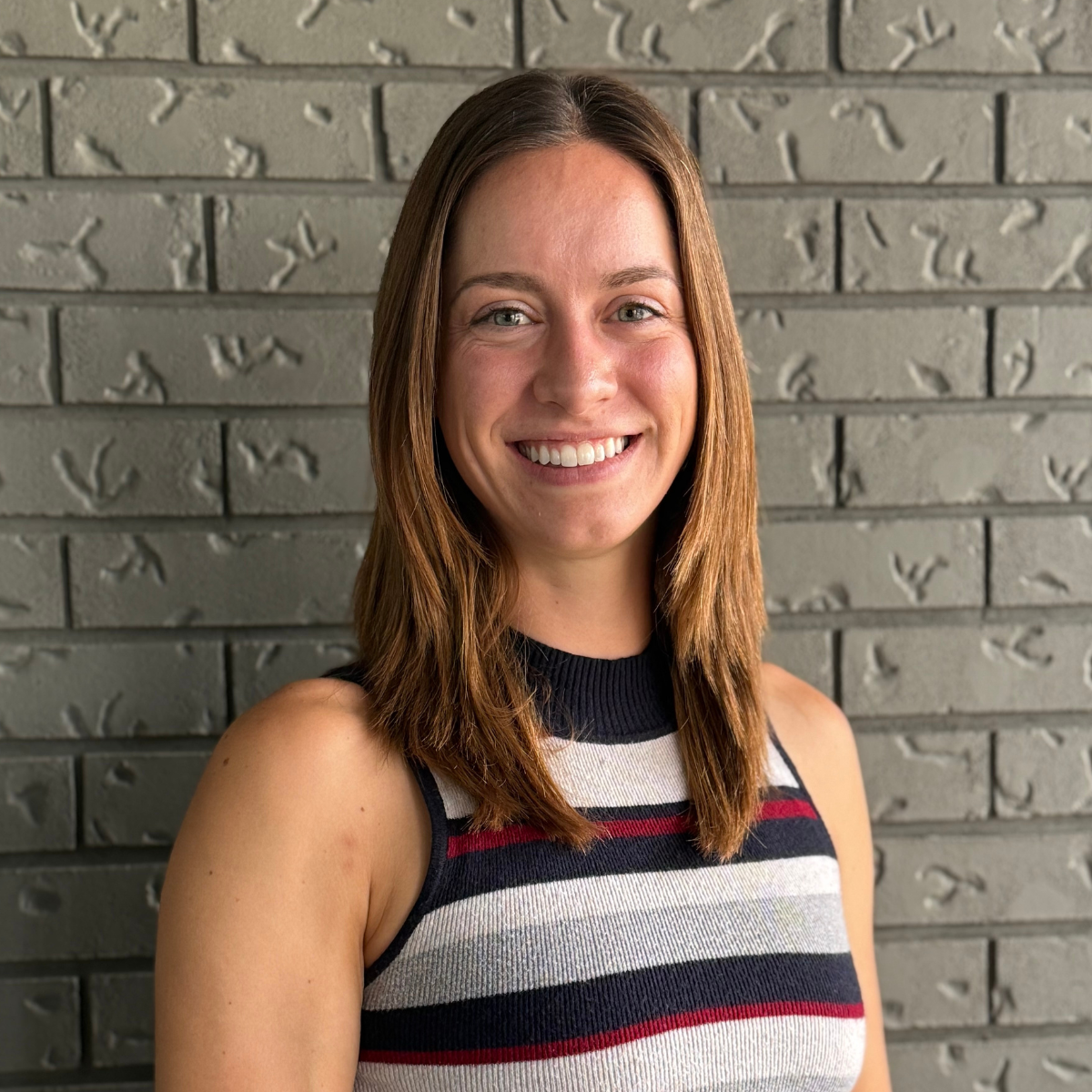 A woman with shoulder-length brown hair, smiling, wearing a sleeveless striped top, standing in front of a gray brick wall.