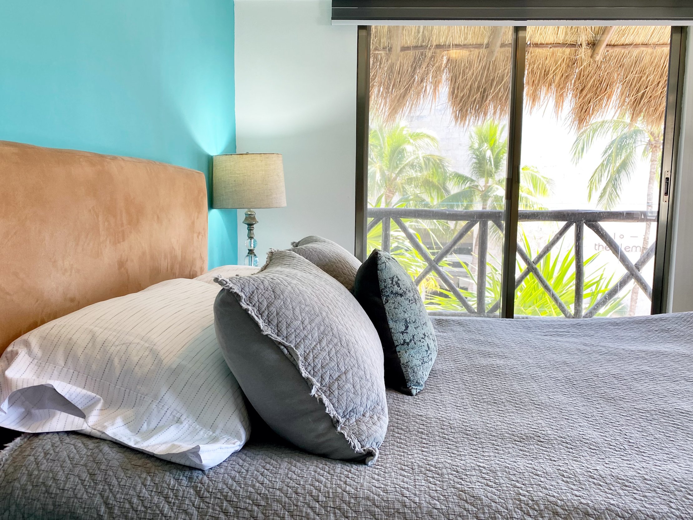 Bedroom with a bed featuring gray and white pillows, blue accent wall, lamp, and sliding glass doors overlooking palm trees.