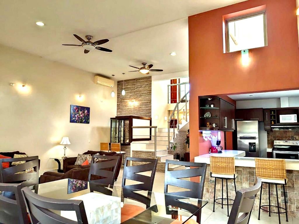 Modern living room and kitchen with glass dining table, brown sofas, ceiling fans, and bar stools. Features a spiral staircase and stainless-steel appliances.
