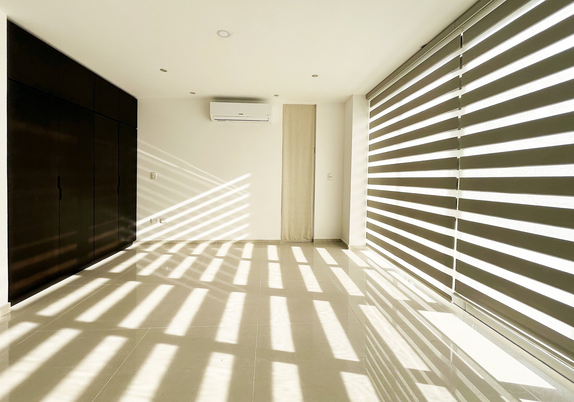 Empty room with sunlight filtering through horizontal blinds, casting shadows on tiled floor. Dark closets on one wall, air conditioner above, and beige accents.
