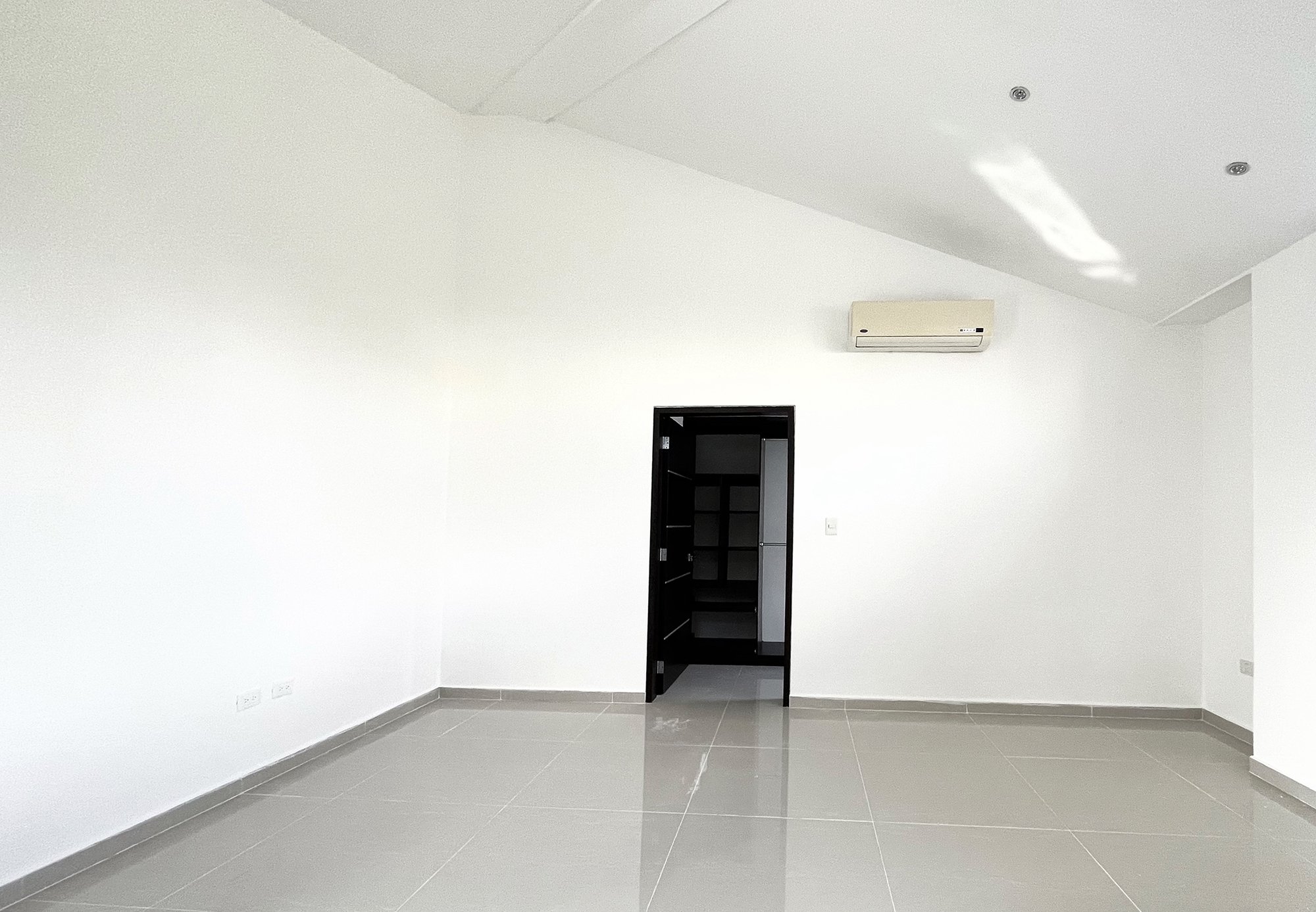 Empty room with white walls, tiled floor, and an air conditioning unit on the wall. A black door leads to a closet area.