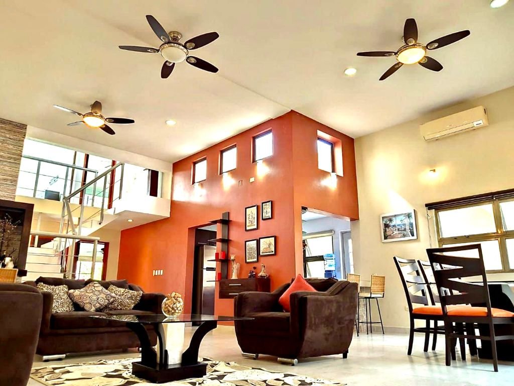 Contemporary living room with brown sofas, glass coffee table, red accent wall, ceiling fans, and dining area.