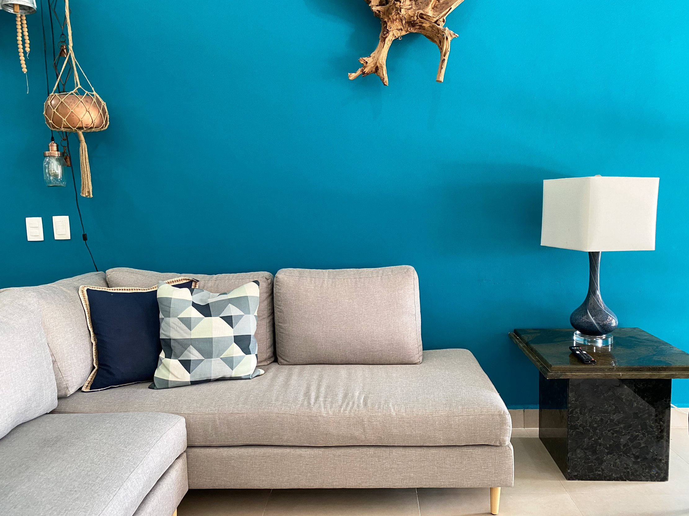 Modern living room with a light gray sectional sofa, blue accent wall, geometric patterned pillow, blue table lamp on a marble side table, and decorative hanging pieces.