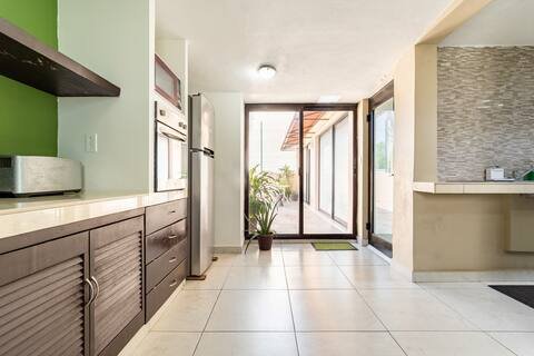 Modern kitchen with white tile floor, wooden cabinets, a stainless steel refrigerator, and a sliding glass door leading to a patio with plants.