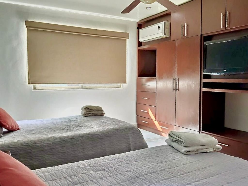 Bedroom with two beds, gray bedspreads, and a brown wardrobe. A TV and air conditioner are mounted on the wall. Folded towels are placed on the beds.
