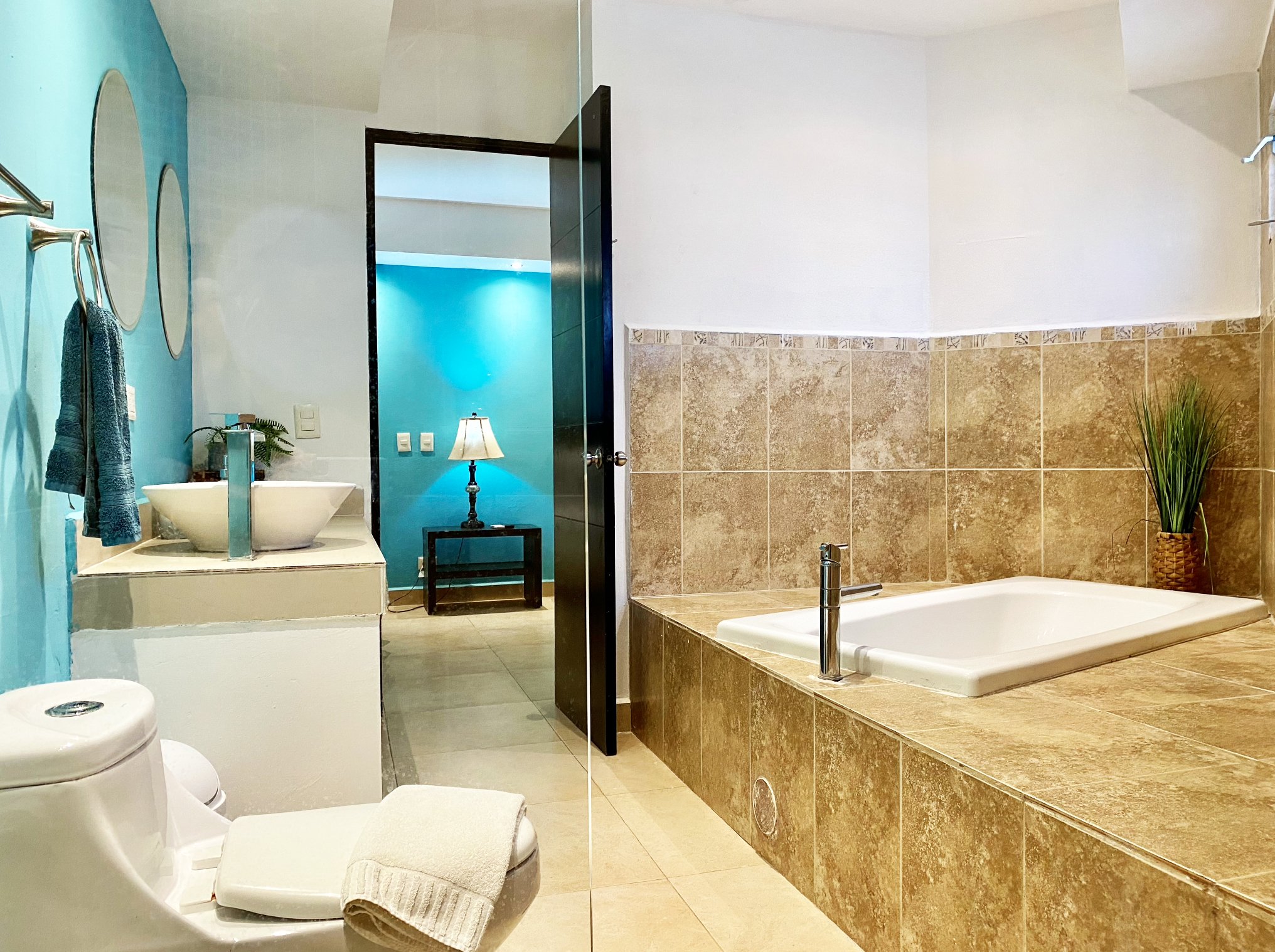 Modern bathroom with blue walls, featuring a white sink, toilet, and large bathtub surrounded by tan tiles. A glass door leads to a room with a lamp on a table.