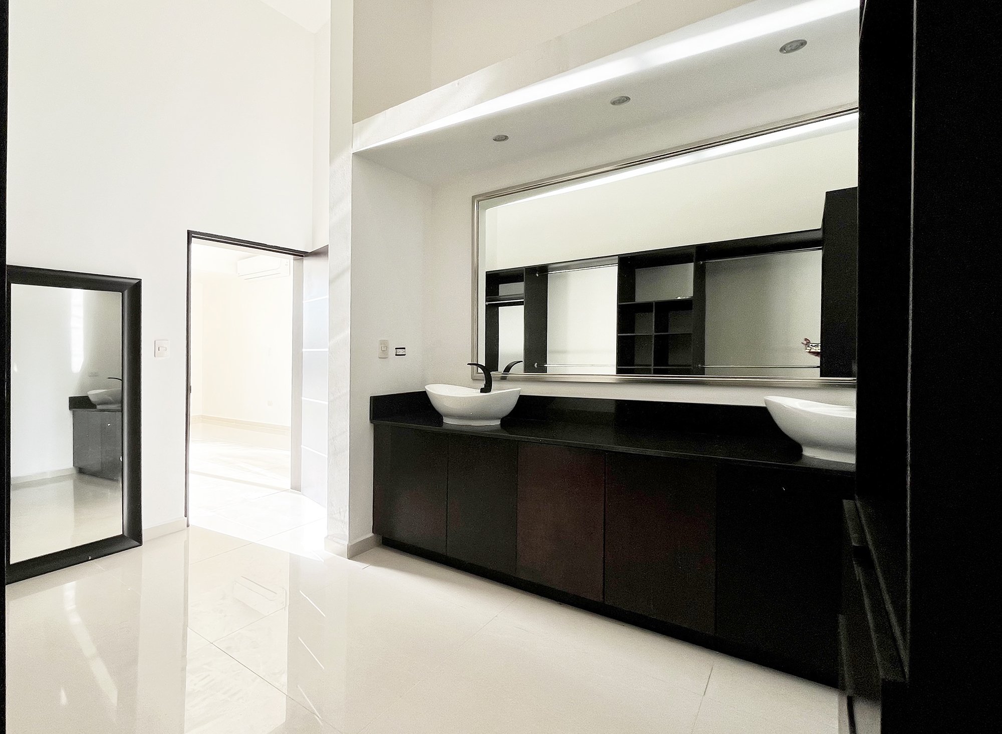 Modern bathroom with double sinks, large mirror, black cabinets, and bright tile flooring. Visible doorway leading to another room.