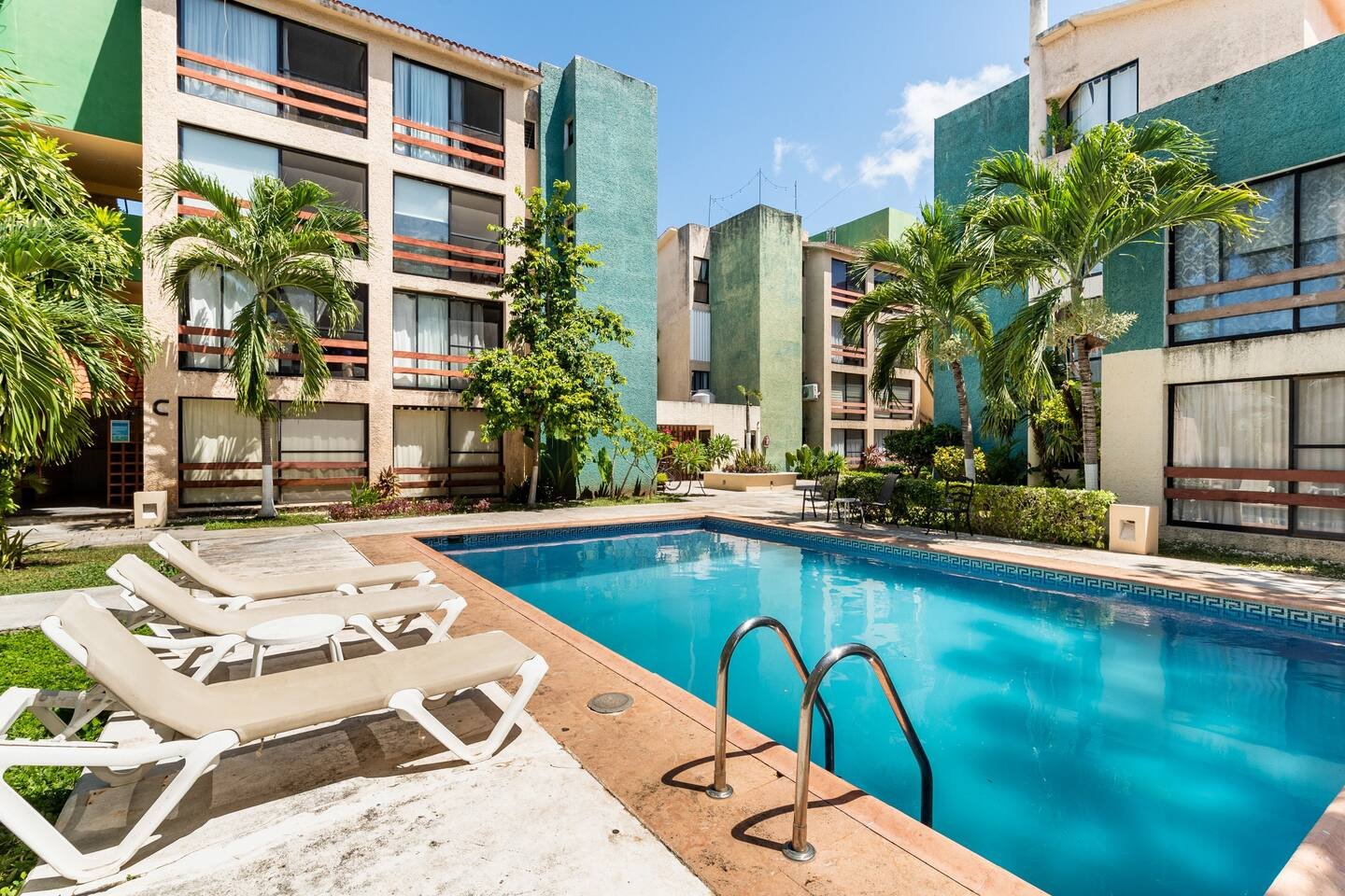 Outdoor swimming pool with lounge chairs and apartment buildings surrounded by palm trees on a sunny day.