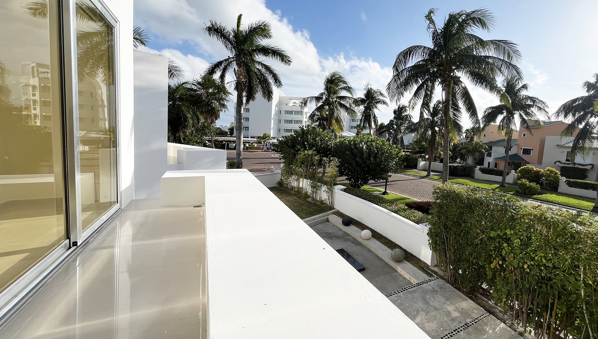 View of a suburban street with palm trees, modern white buildings, and landscaped gardens under a blue sky with clouds.