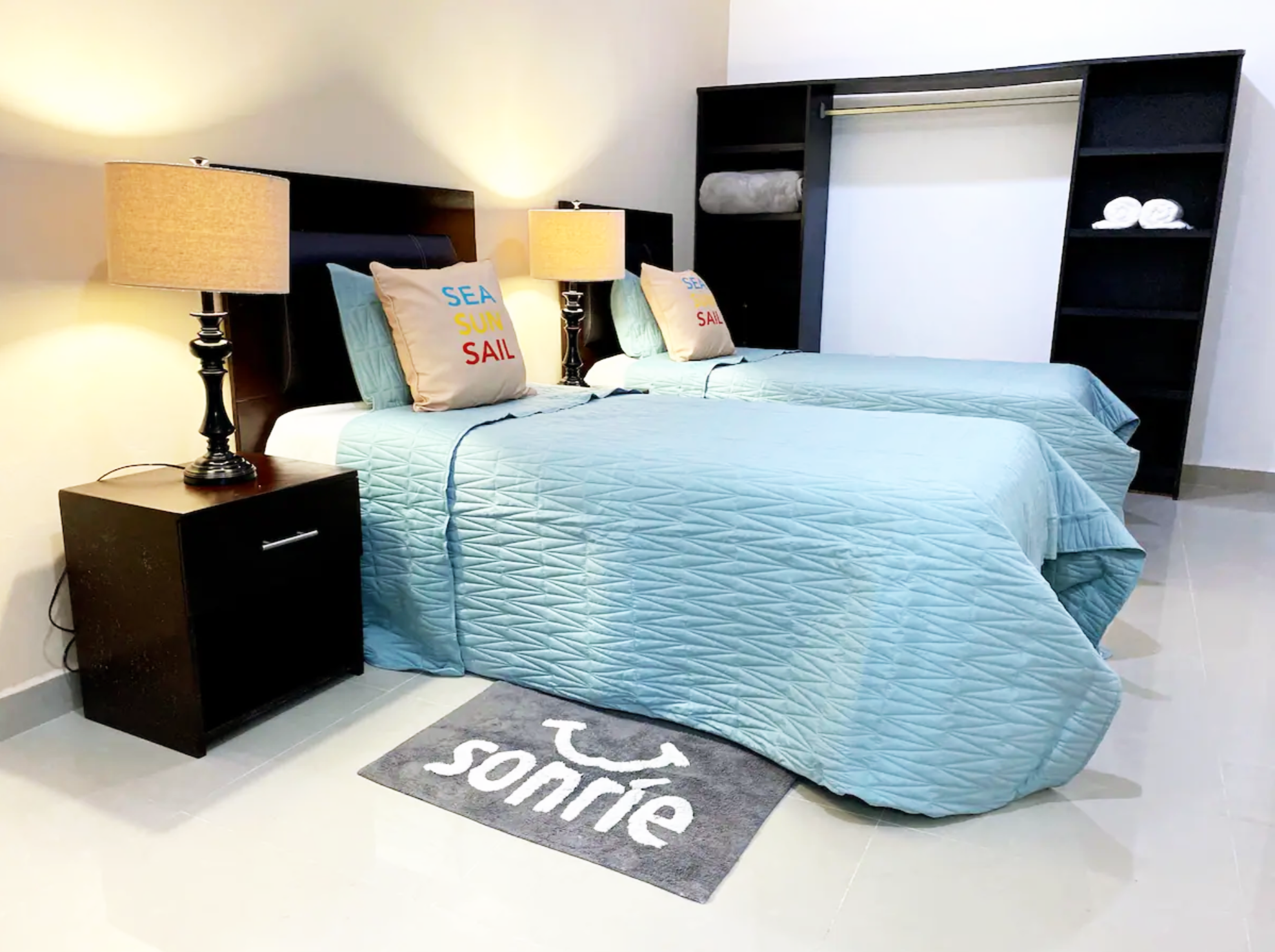 Twin bedroom with light blue bedding, two bedside lamps, pillows with 'Sea, Sun, Sail' text, dark wood nightstand, empty closet, and a rug with 'sonríe' text.