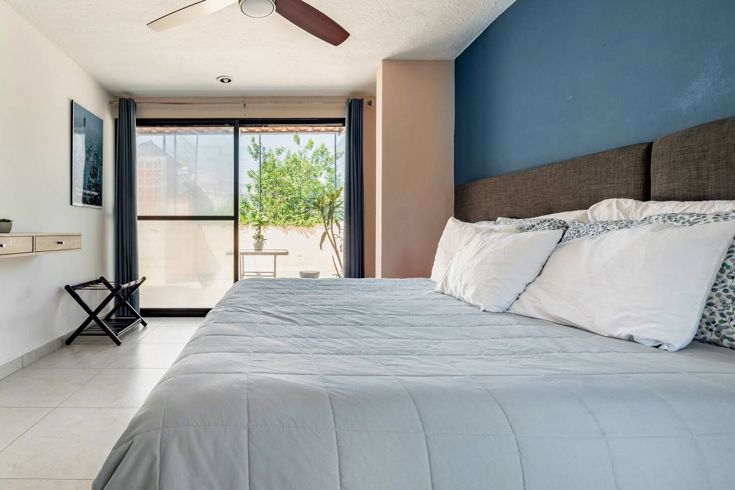 Modern bedroom with blue accent wall, queen-sized bed with white bedding, large sliding glass door leading to a balcony with plants, ceiling fan, and wall-mounted desk.
