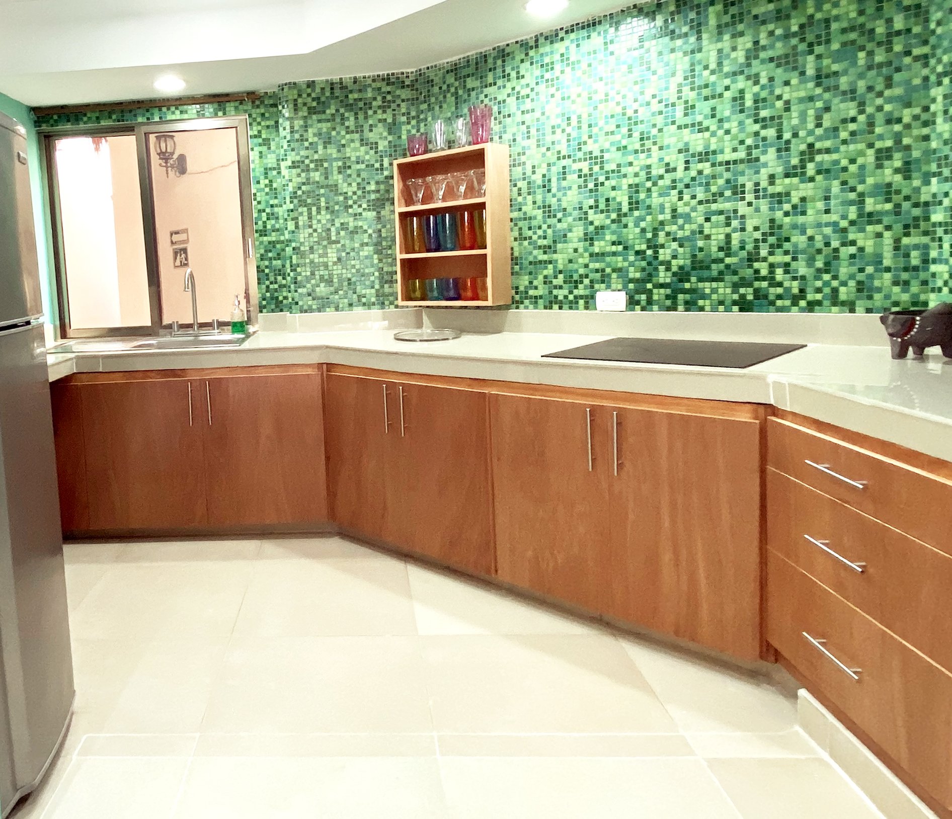 Modern kitchen with green mosaic backsplash, wooden cabinets, stainless steel fridge, and colorful glassware on a shelf.