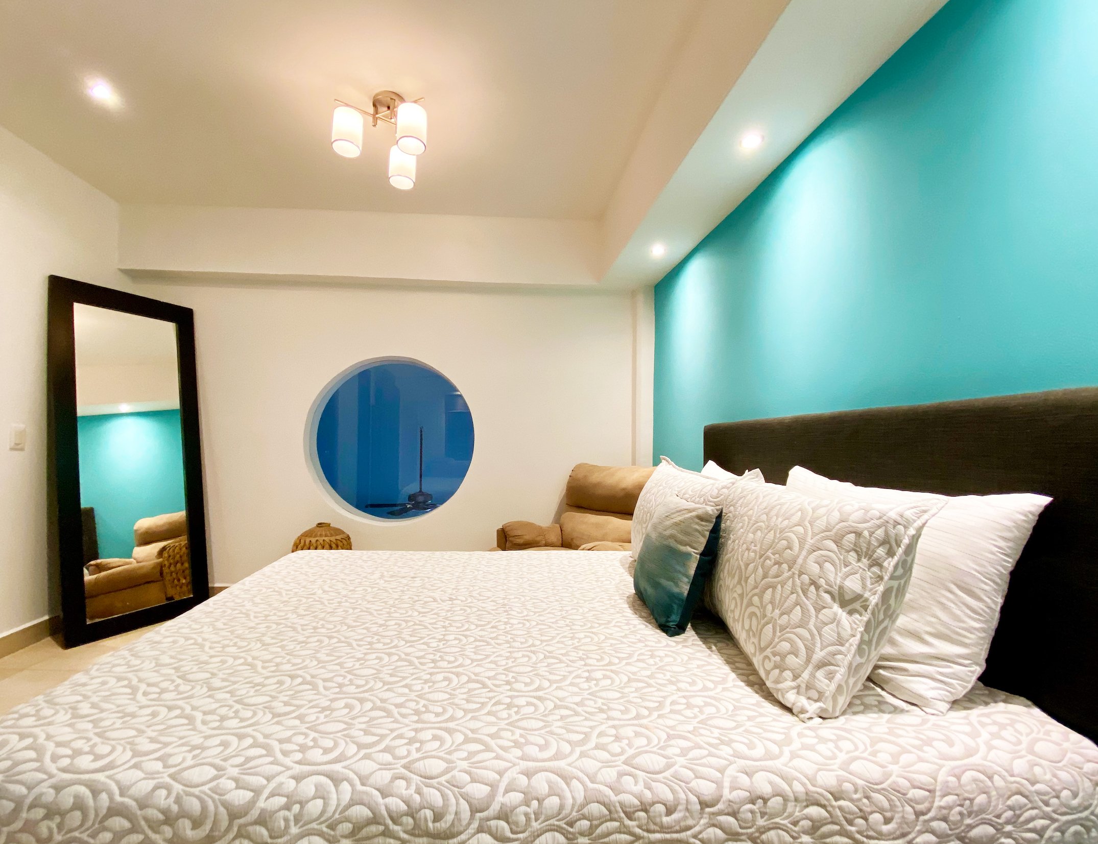 Modern bedroom with a circular window, a queen-sized bed with patterned bedding, decorative pillows, a large mirror, and an upholstered chair. Bright teal accent wall and ceiling lights enhance the space.