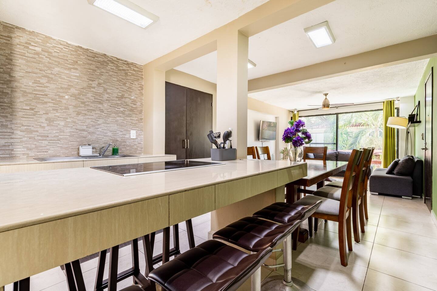 Modern open-concept kitchen and dining area with a beige countertop, bar stools, dining table with chairs, purple flowers, and a living area with a sofa and a TV.