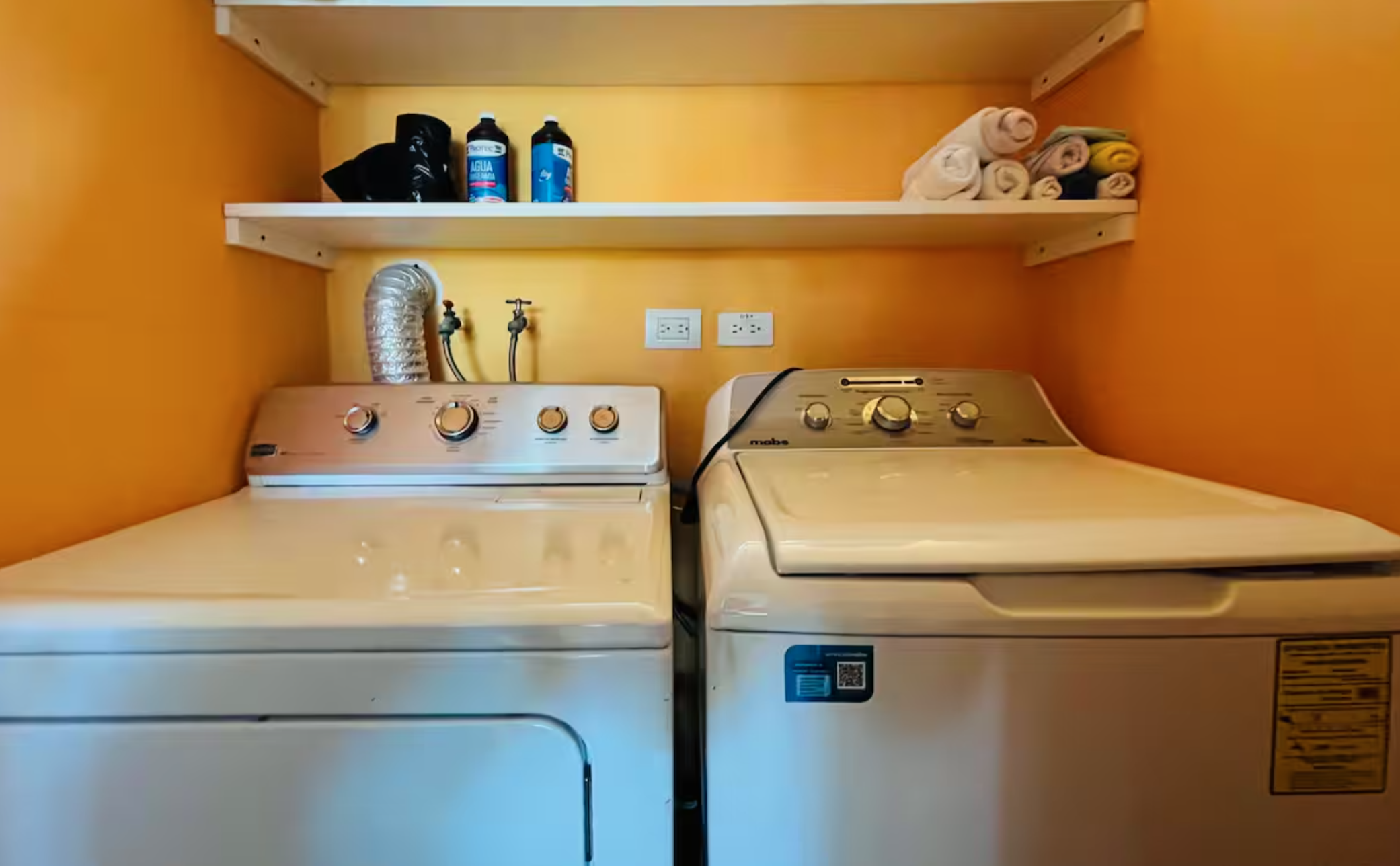 Laundry room with a washer and dryer, orange walls, shelves holding detergent and towels.