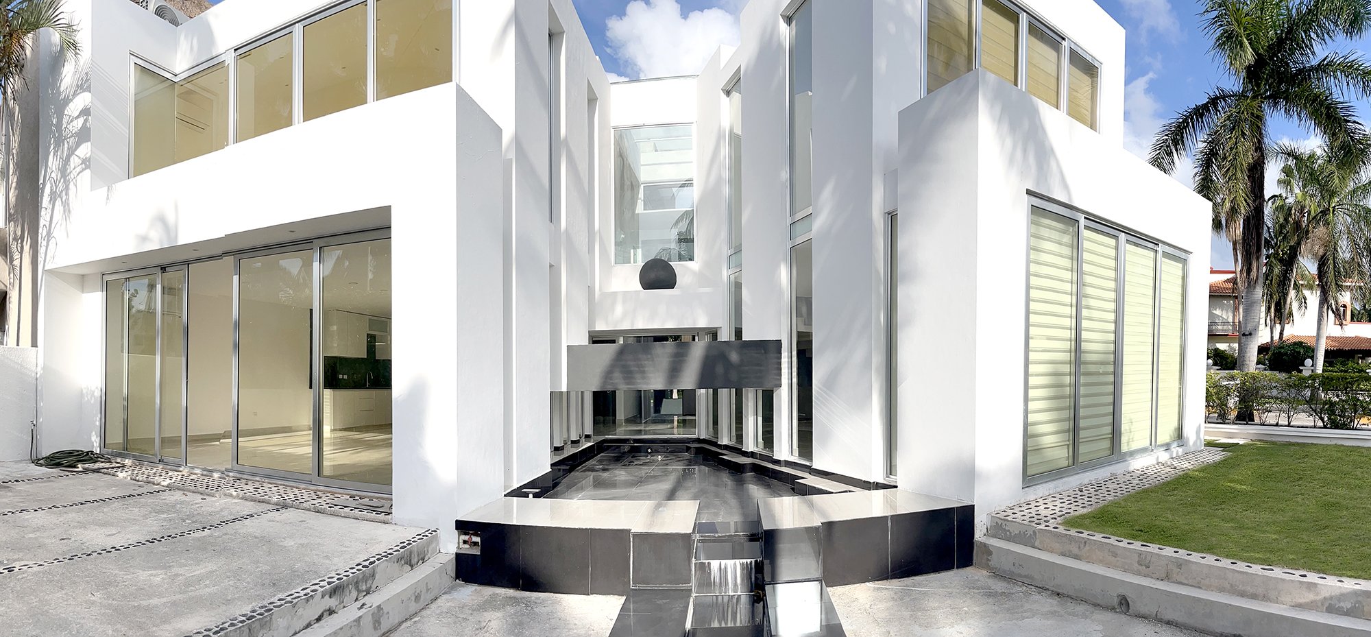 Modern white house with large windows and a central courtyard, featuring a small black-tiled water feature. Palm trees are visible in the background.