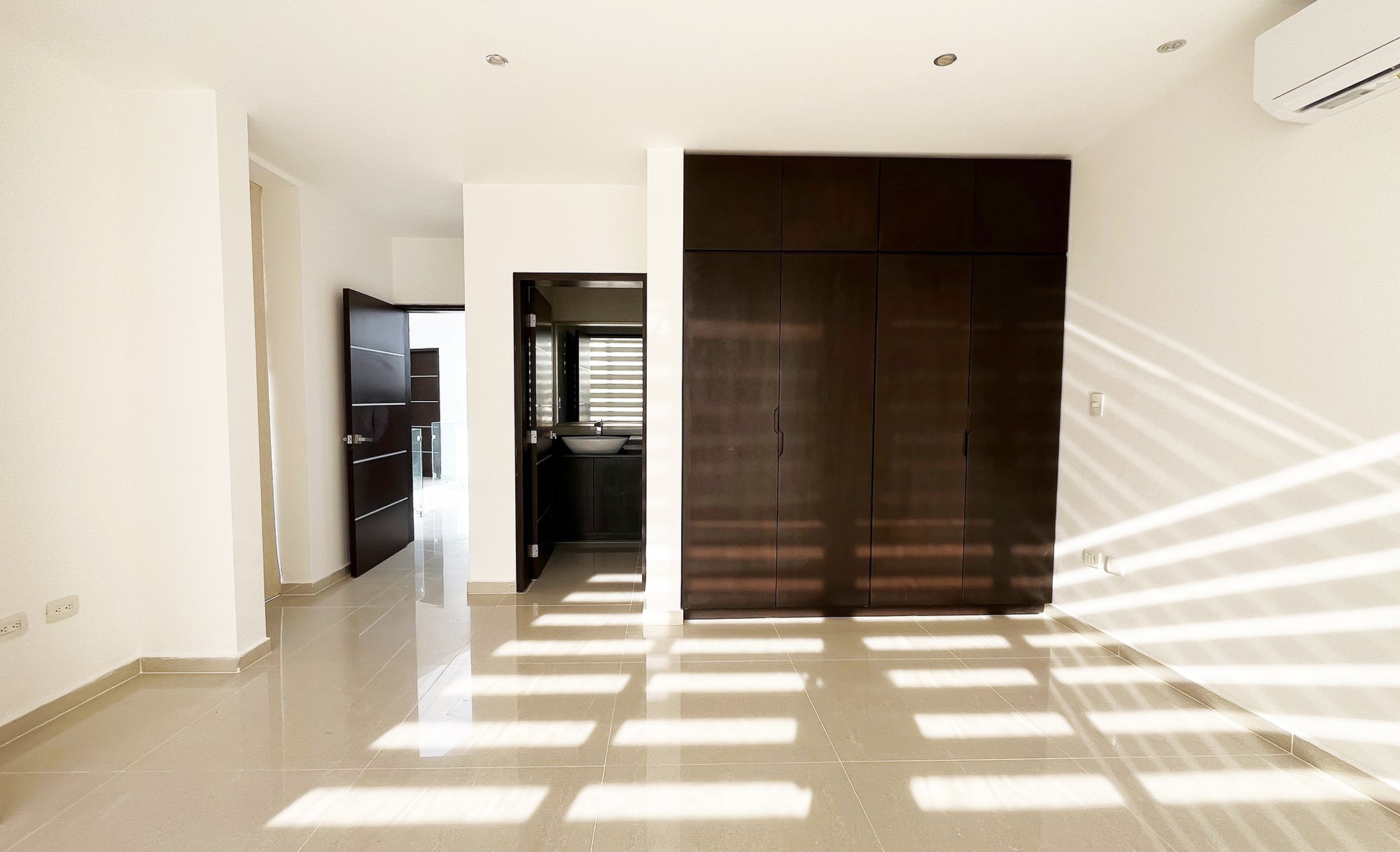 Empty room with beige tile flooring, dark wooden wardrobe, open door, sunlight creating striped patterns, and wall-mounted air conditioner.