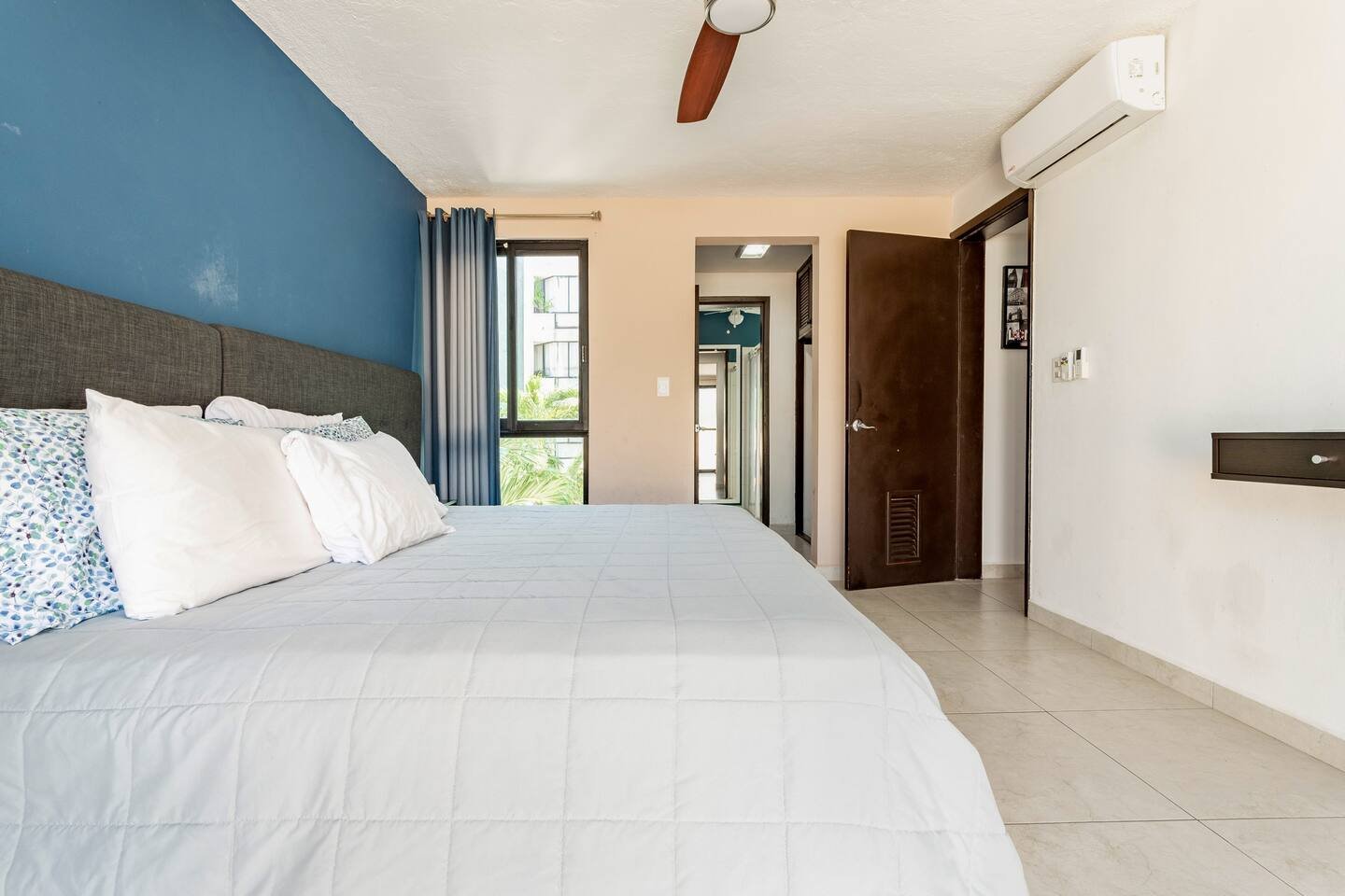 Bedroom with blue accent wall, bed with white bedding, window with blue curtains, ceiling fan, open door, and air conditioning unit.