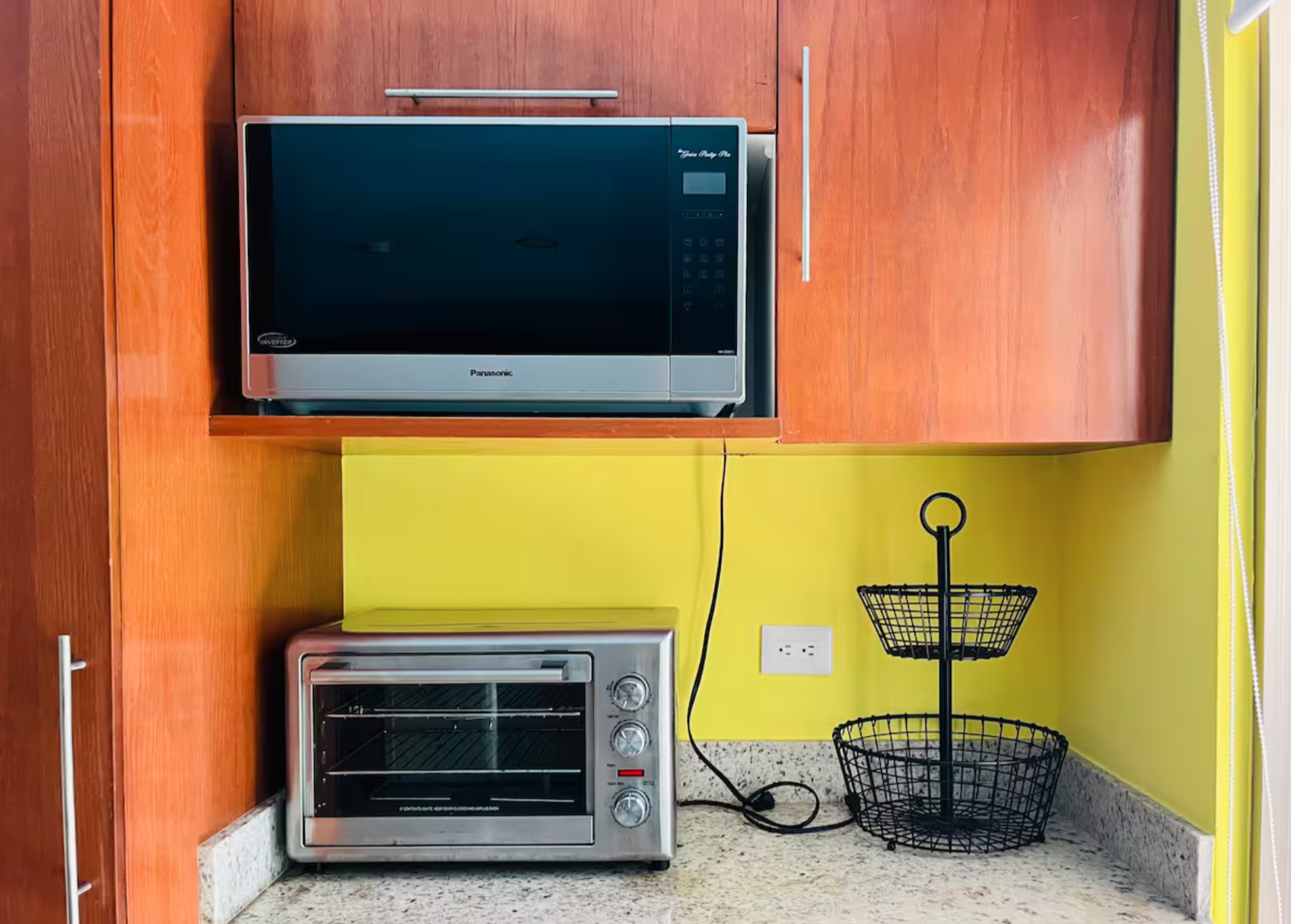 Kitchen with a microwave mounted in a wooden cabinet, a toaster oven on a countertop, and a two-tiered wire fruit basket, against a yellow wall.
