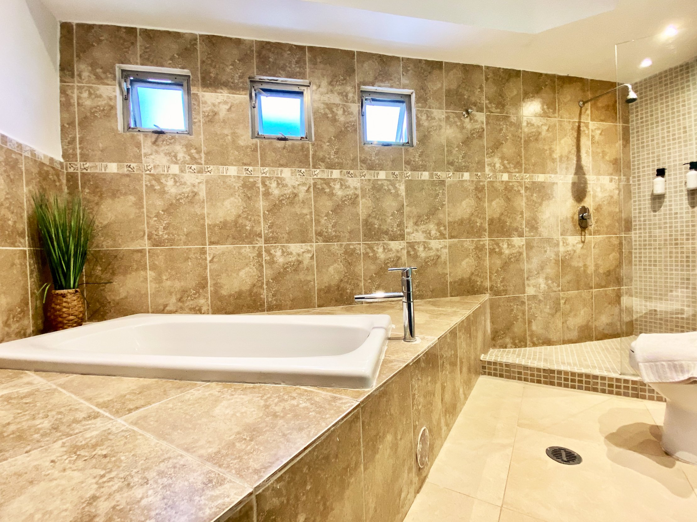 Modern bathroom featuring a bathtub, shower area, and brown tiled walls. Three small windows above the tub and a potted plant provide a decorative touch. The shower has a wall-mounted showerhead, and the floor is tiled.