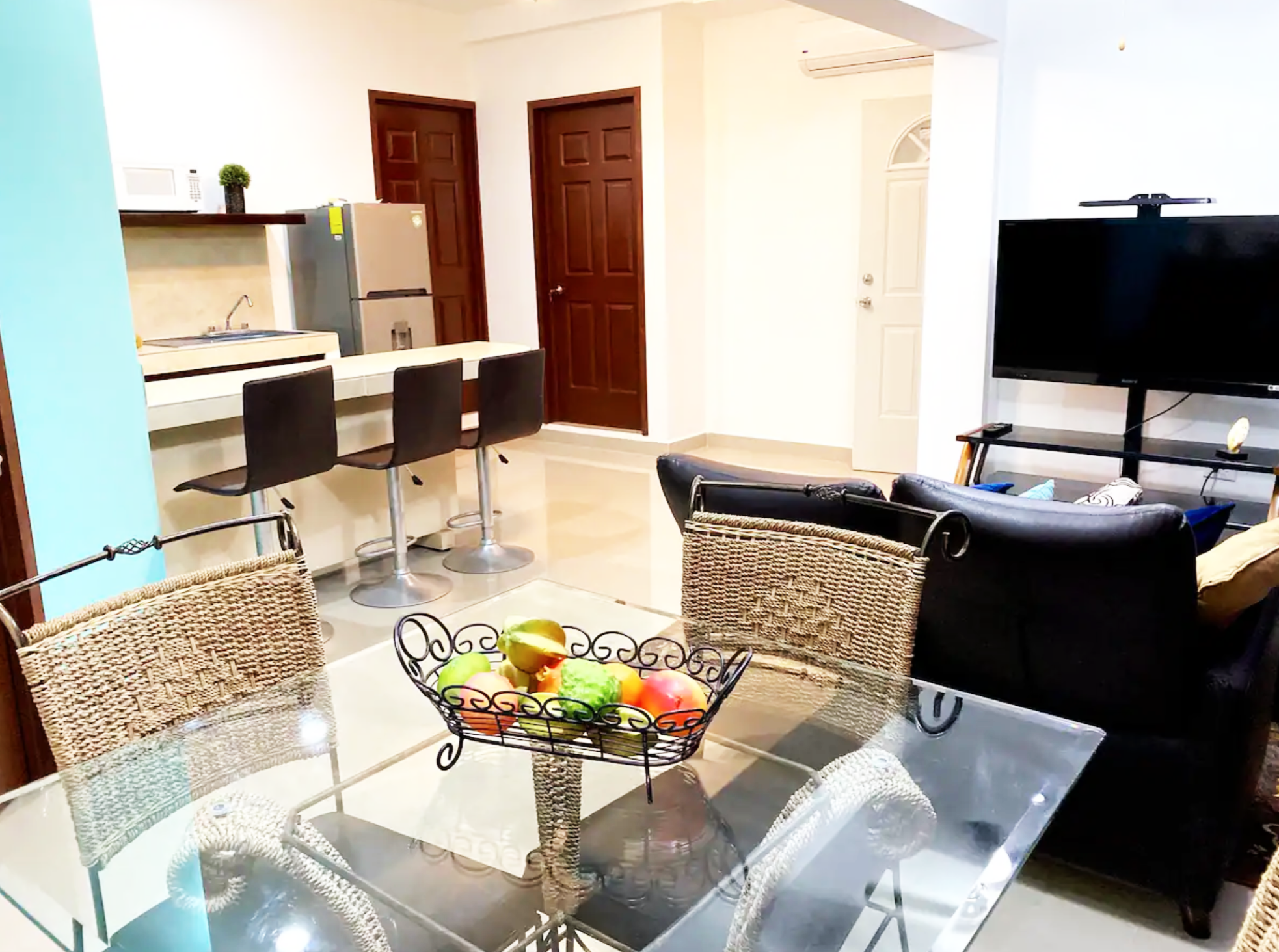 A modern kitchen and living area with a glass dining table, wicker chairs, a wire fruit basket on the table, two bar stools at a counter, a refrigerator, a TV, a couch, and wooden doors.