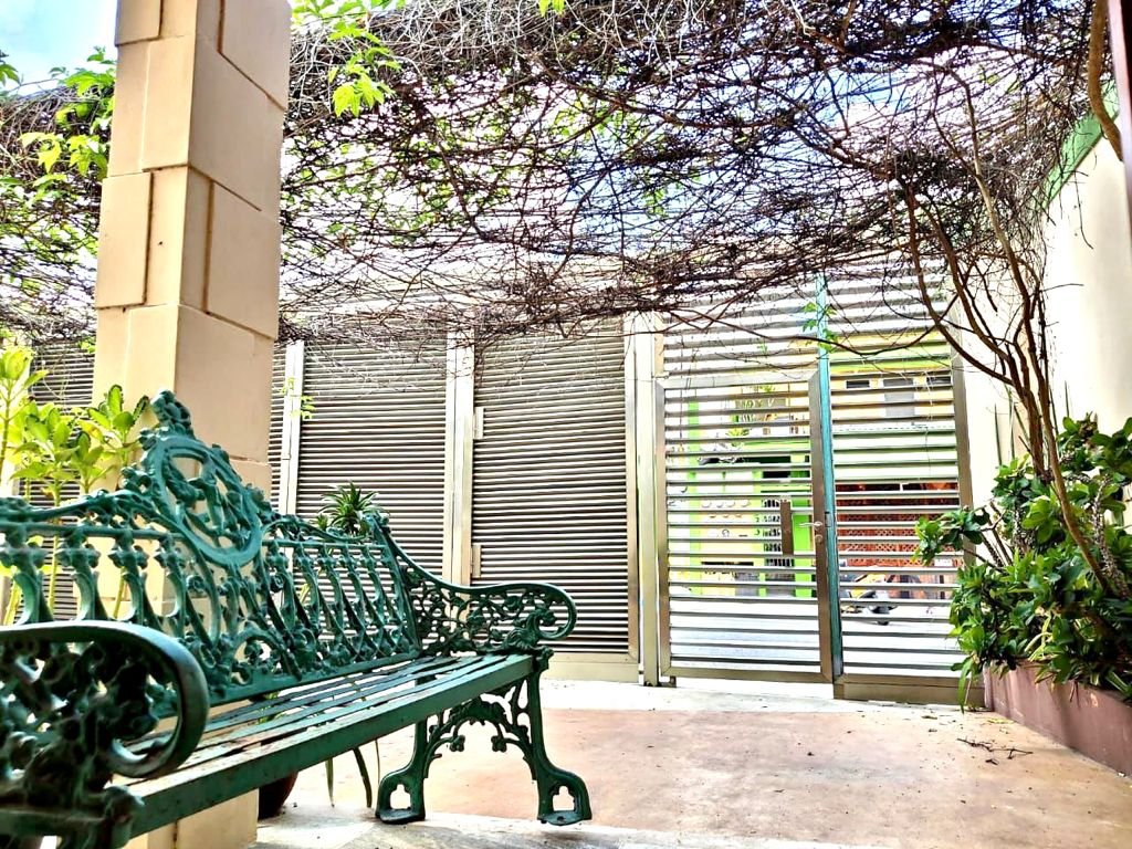 Outdoor area with an ornate metal bench, surrounded by greenery and a trellis covered in vines.
