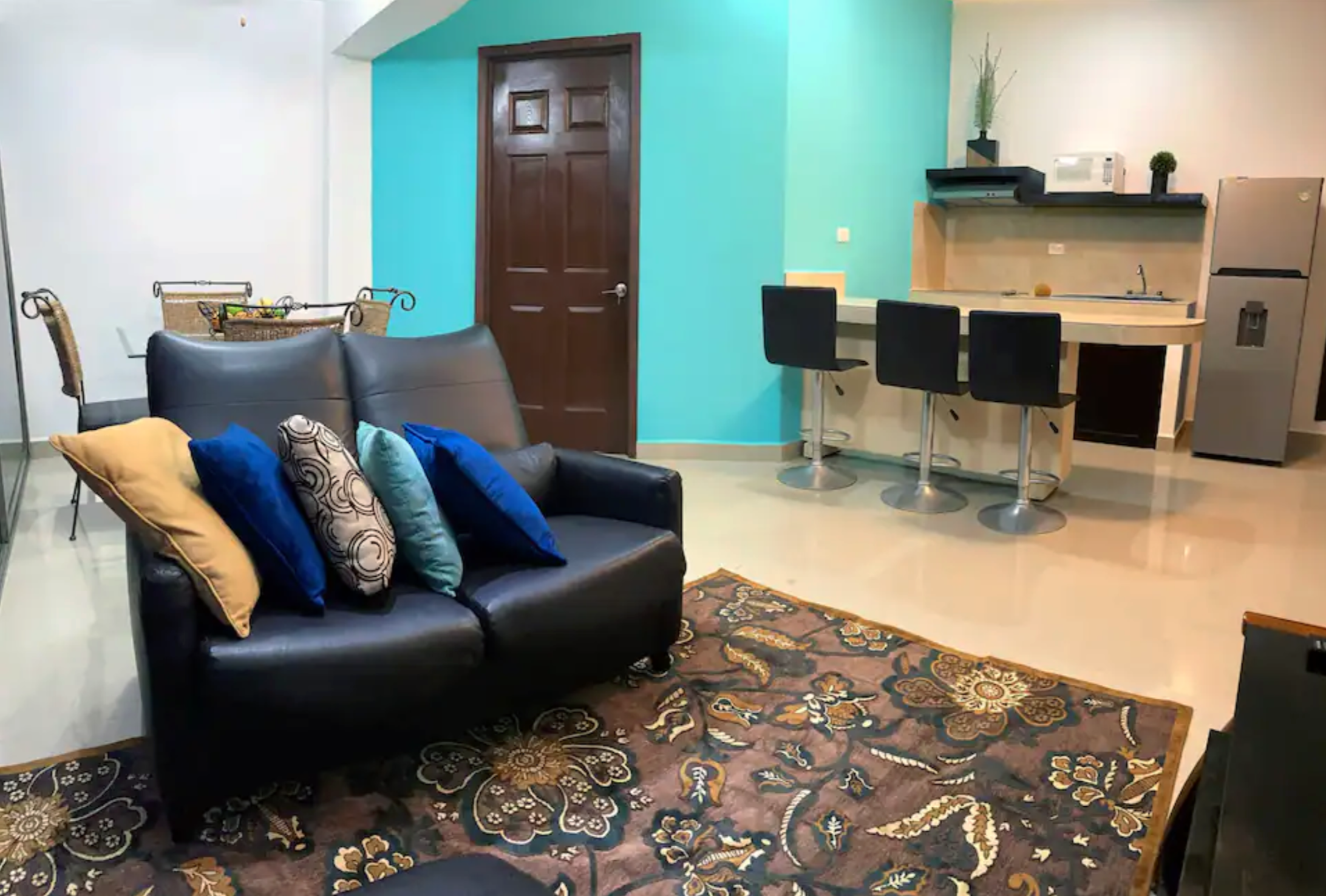 Modern living room with a black sofa, colorful cushions, patterned rug, bar counter with stools, kitchen area, and dining table in the background.