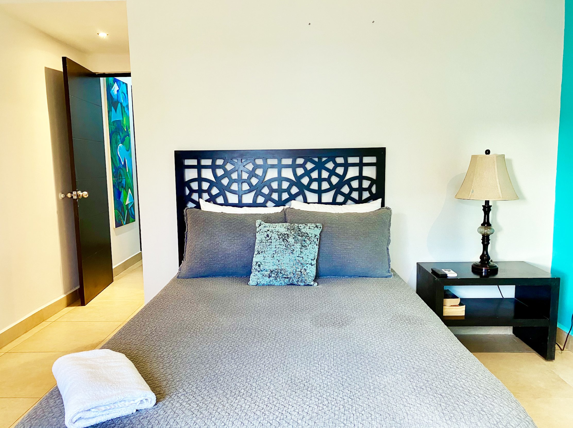 Bedroom with a bed featuring gray bedding and pillows, a decorative headboard, a lamp on a nightstand, an open door, and a towel folded at the foot of the bed.