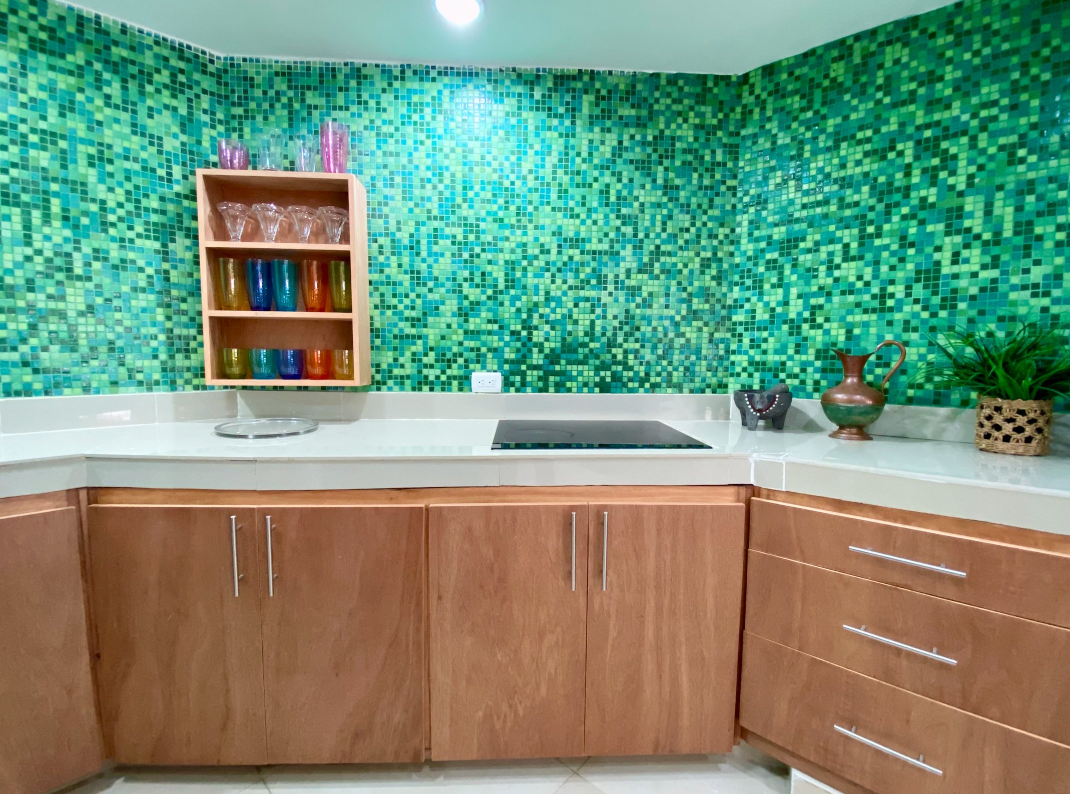 Kitchen with wood cabinets, green mosaic tile backsplash, colorful glassware on a shelf, stovetop, and decorative items on the counter.