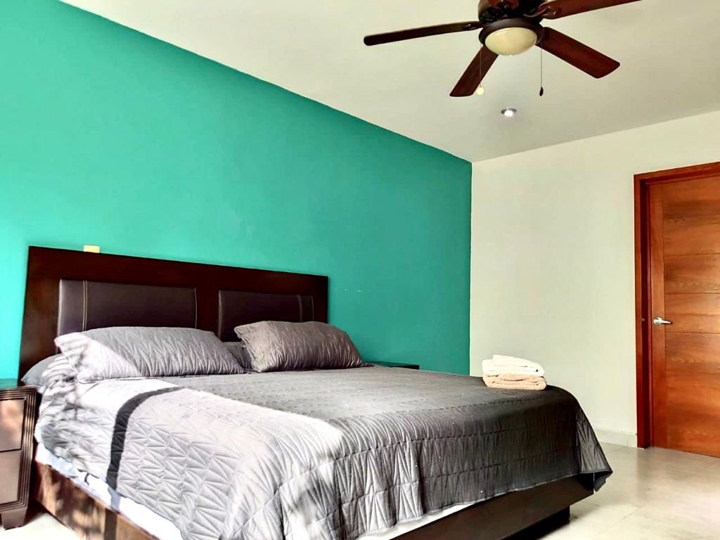 Bedroom with teal accent wall, large bed with gray bedding, ceiling fan, wooden door, and folded towels on bed.