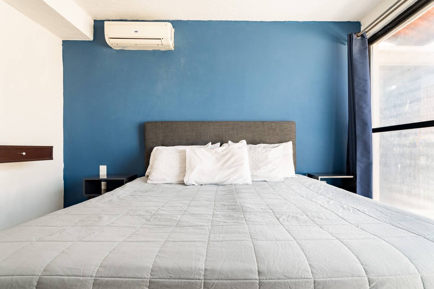 Minimalist bedroom with a gray bed against a blue wall, white bedding, and an air conditioner above.