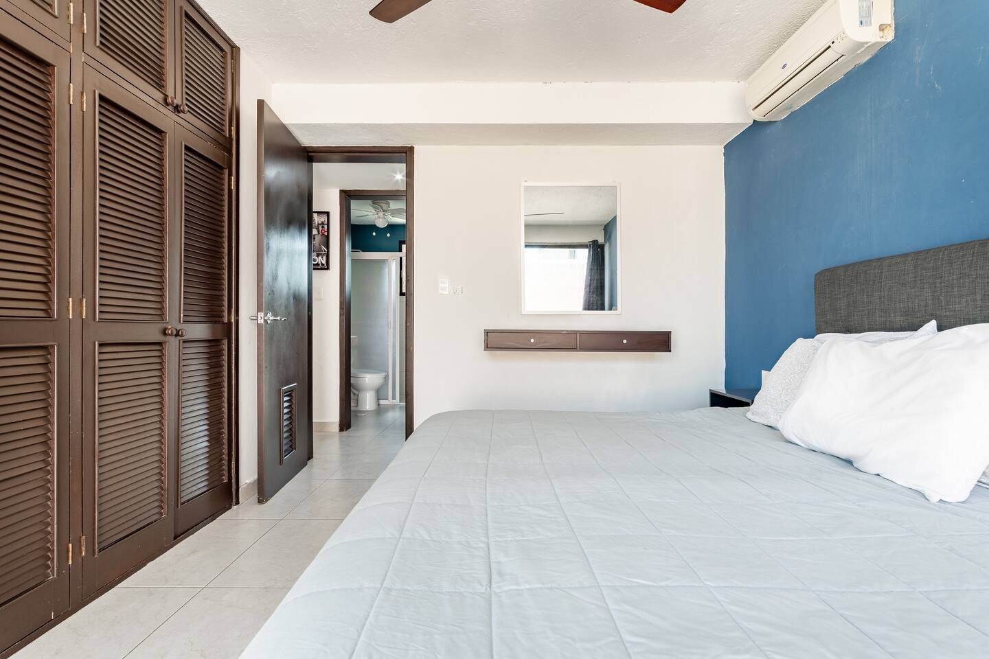 Modern bedroom with a bed, blue accent wall, ceiling fan, wooden closet, and an open door leading to a bathroom.