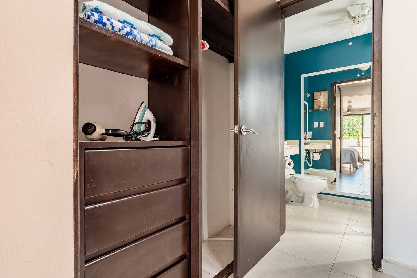 Open closet and view of bathroom with wooden shelving, towels, hairdryer, and iron; bathroom with blue walls, mirror, toilet, and visible part of a bedroom.