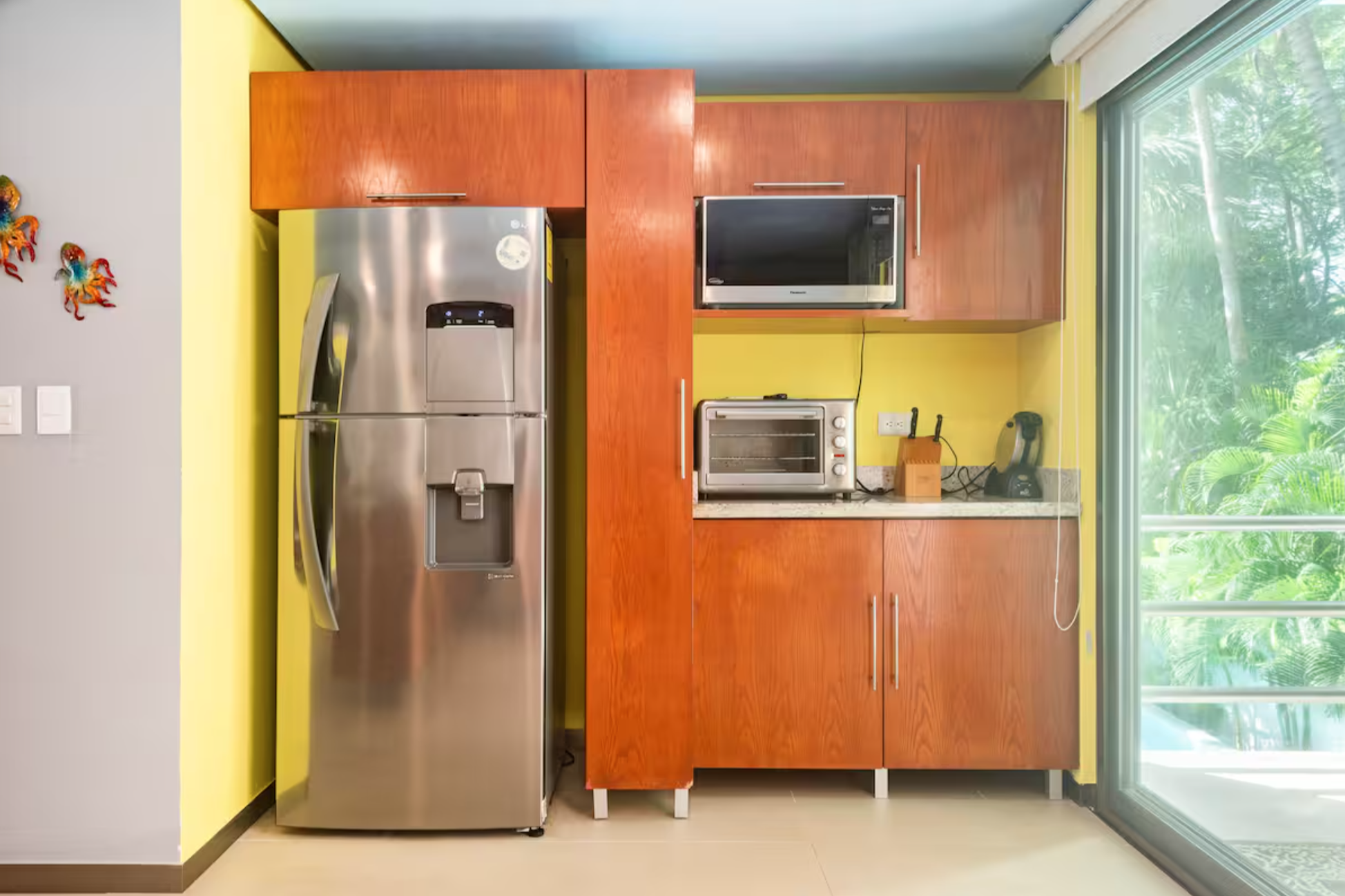 Modern kitchen with stainless steel refrigerator, wooden cabinets, microwave, toaster oven, knife block, and blender, beside a large window with a view of greenery.