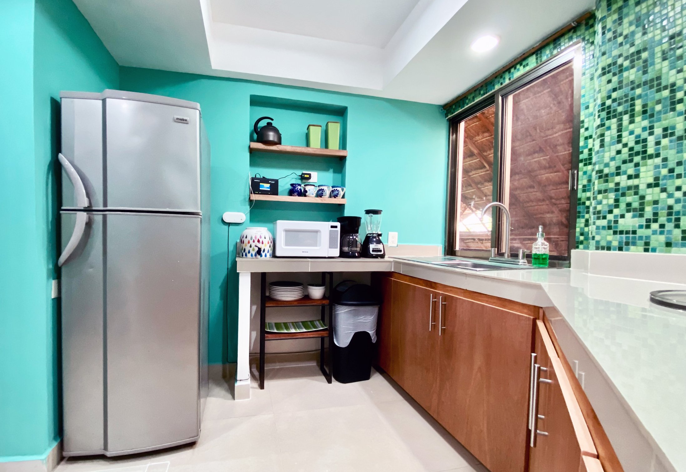 Modern kitchen with teal walls, featuring a silver refrigerator, microwave, coffee maker, blender, and shelves with dishes. Large window with green mosaic tiles above sink.