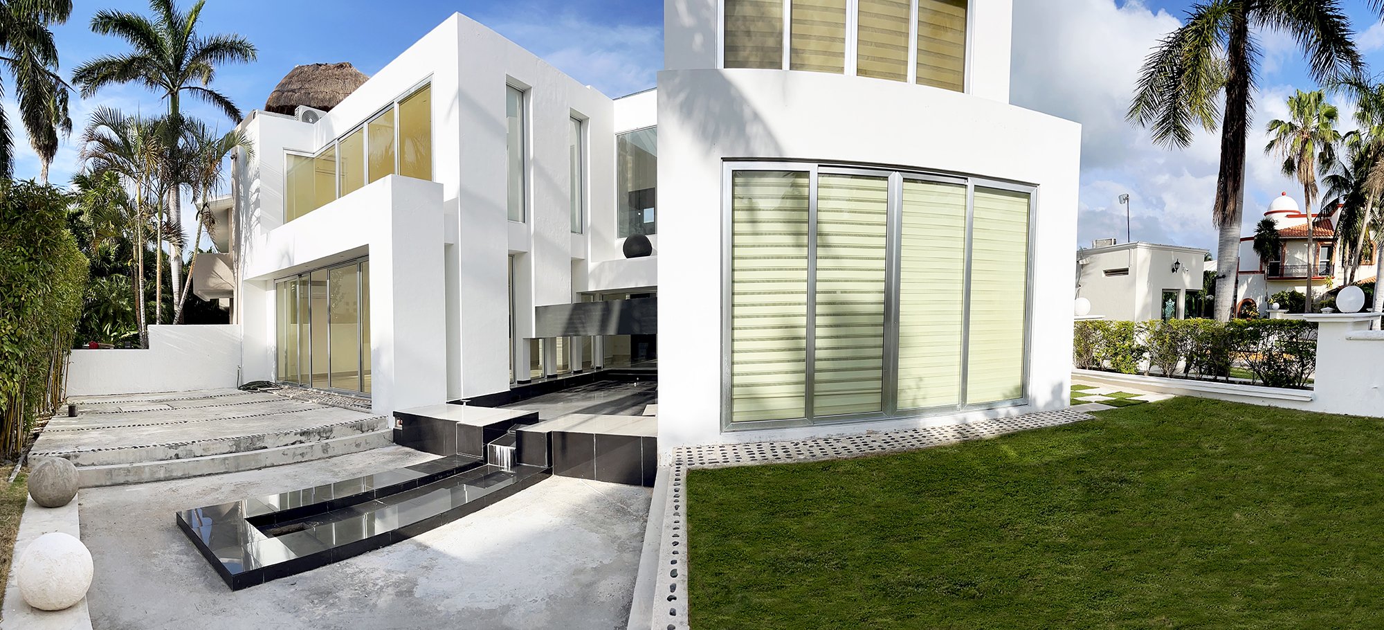Modern white house with large windows and a minimalist design, surrounded by tropical plants and palm trees. The exterior features a geometric black water feature and a neatly trimmed lawn.