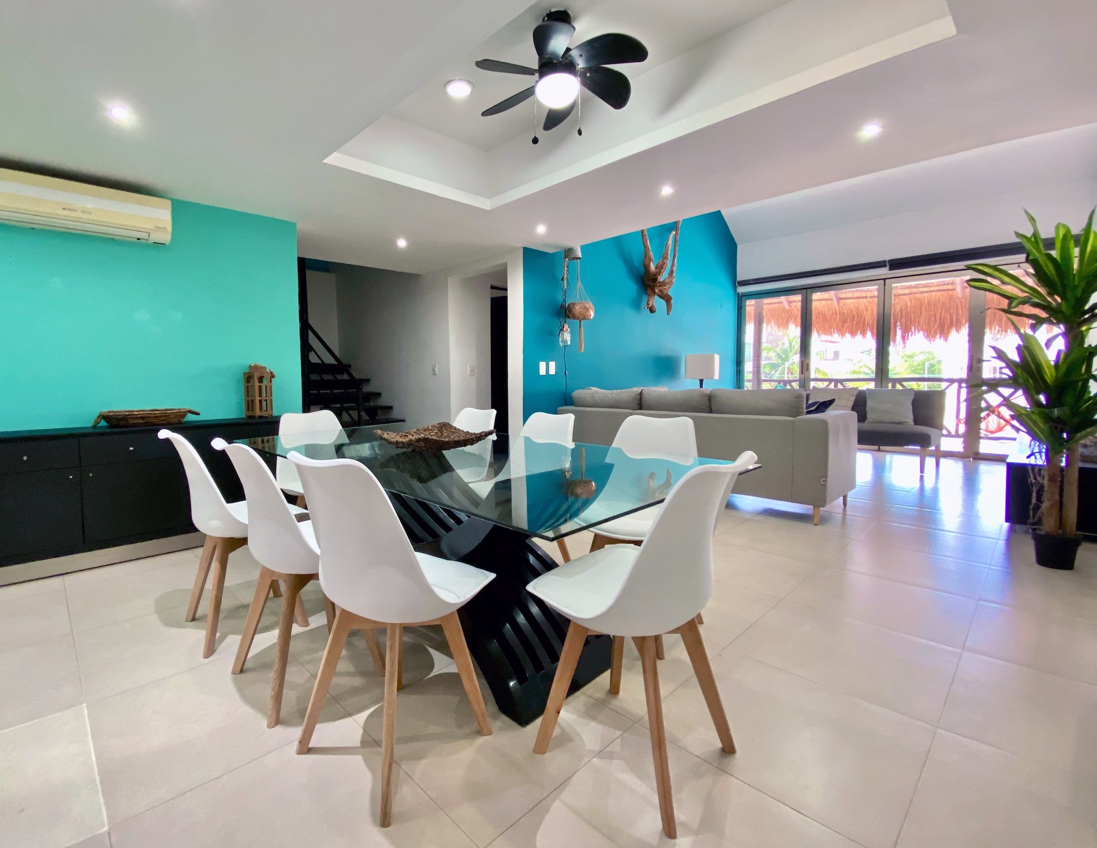 Modern dining and living room with white chairs, glass table, gray couch, teal accent wall, indoor plants, ceiling fan, and large windows.
